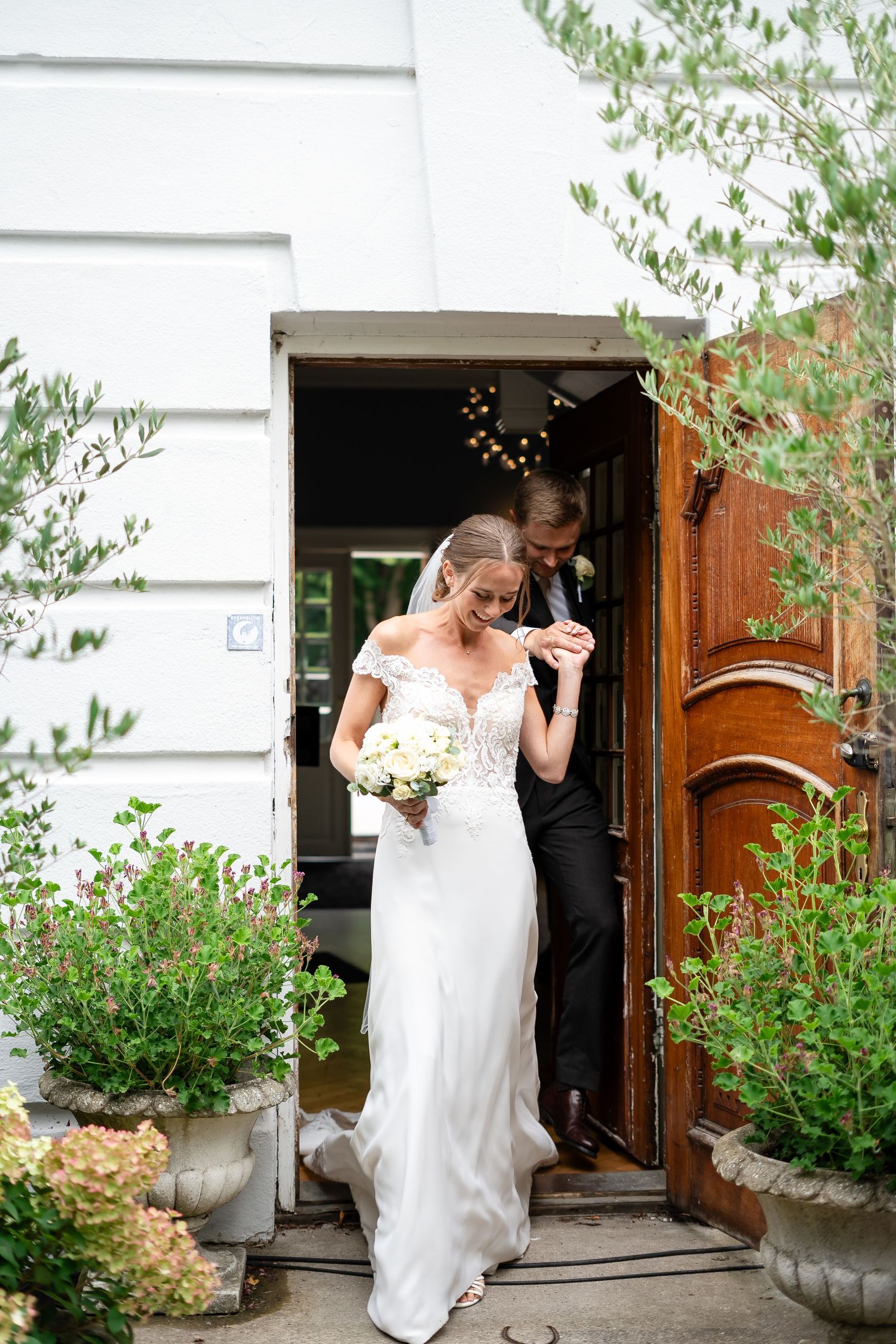 Bride in white lace gown holding bouquet exits wooden doors with groom at elegant venue surrounded by greenery.