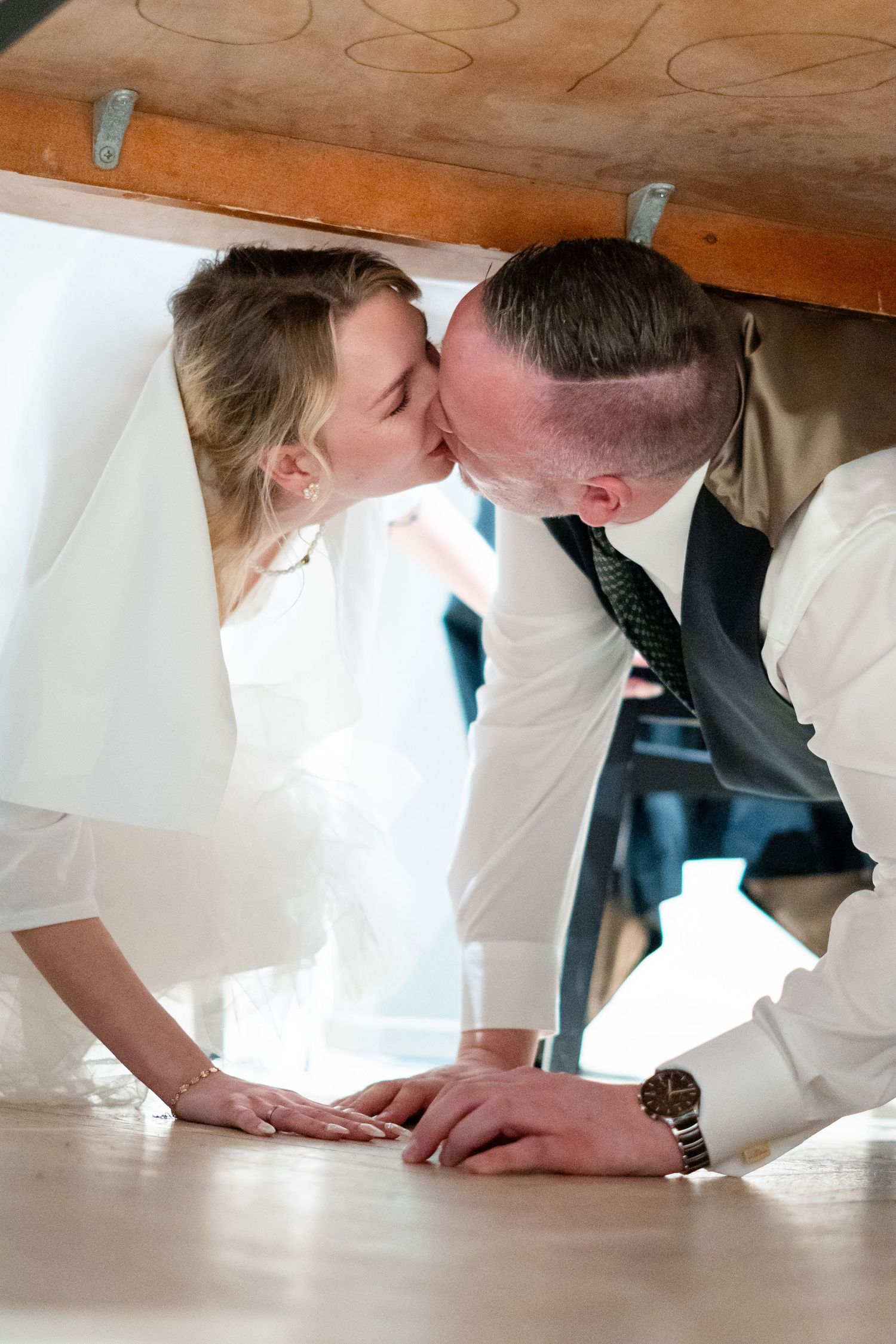 Bride and groom sharing a romantic kiss under a wooden table on their wedding day.