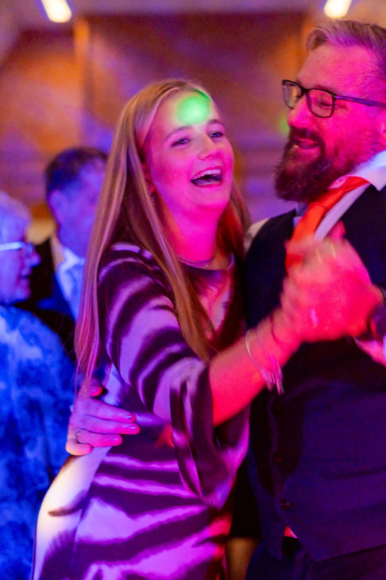 A joyful couple dancing together at a festive event, illuminated by colorful purple and pink lights.