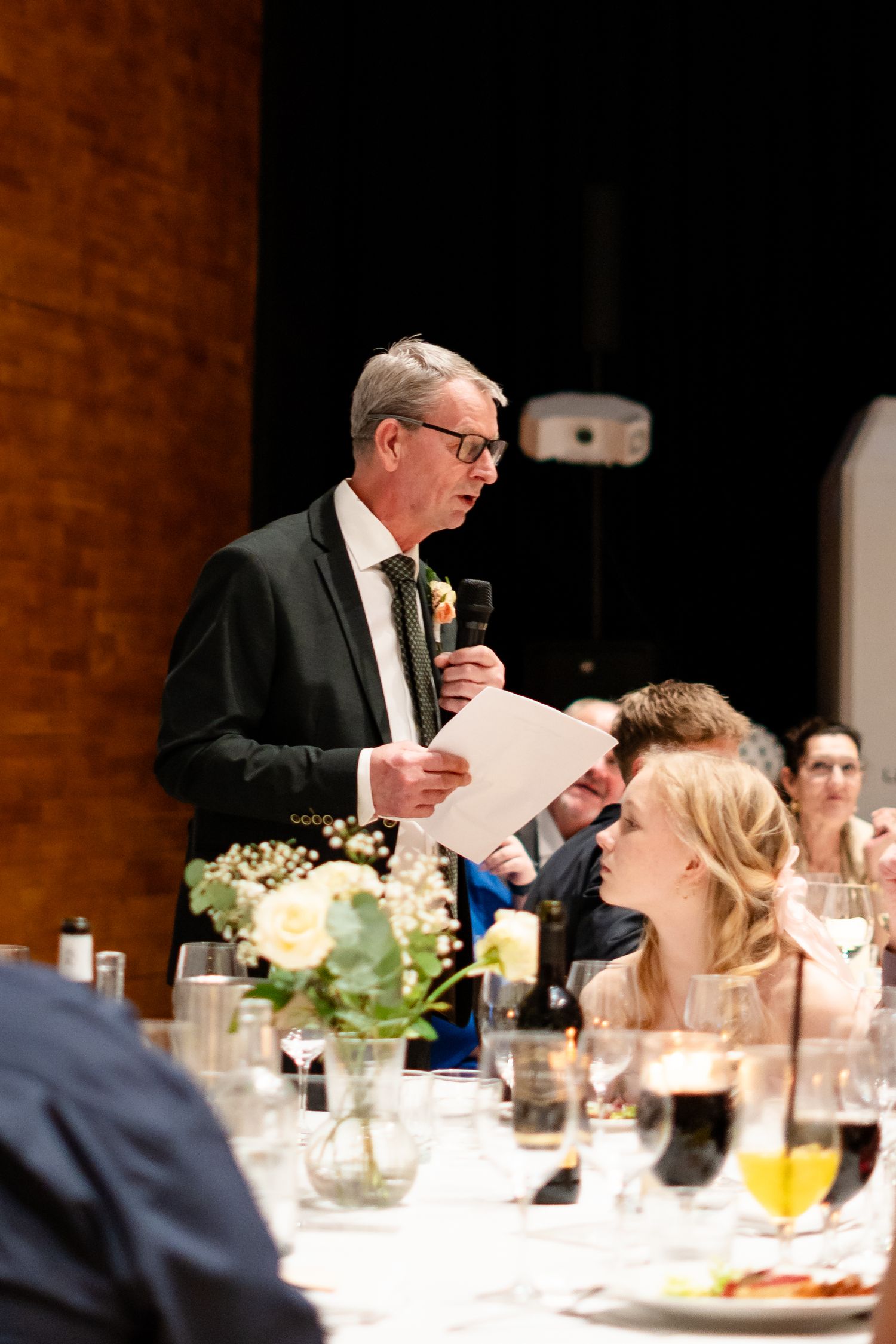Man in suit giving a speech at a wedding reception dinner, holding microphone and papers at an elegant table.