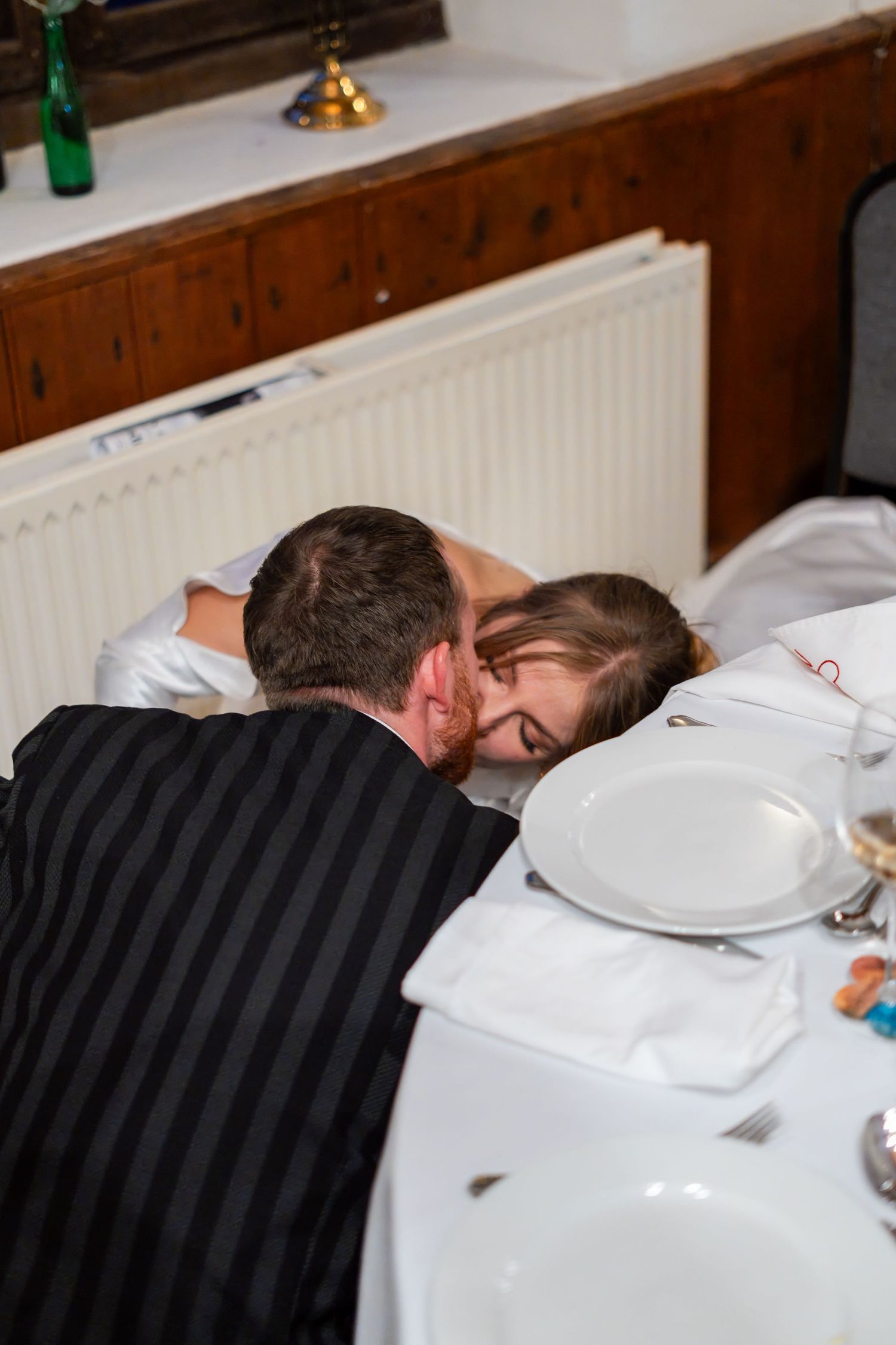 Couple sharing a romantic kiss at a wedding reception dinner table with white tablecloth and plates.