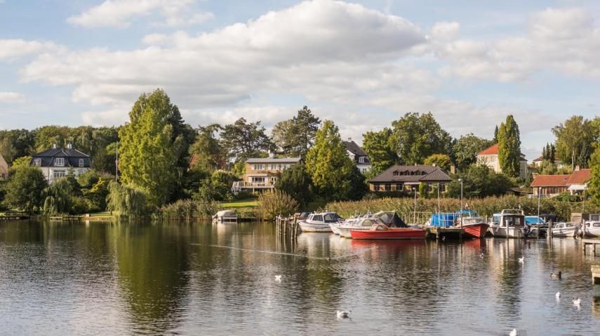 Panoramic view of a waterfront with boats docked along a tree-lined shore on a sunny day.