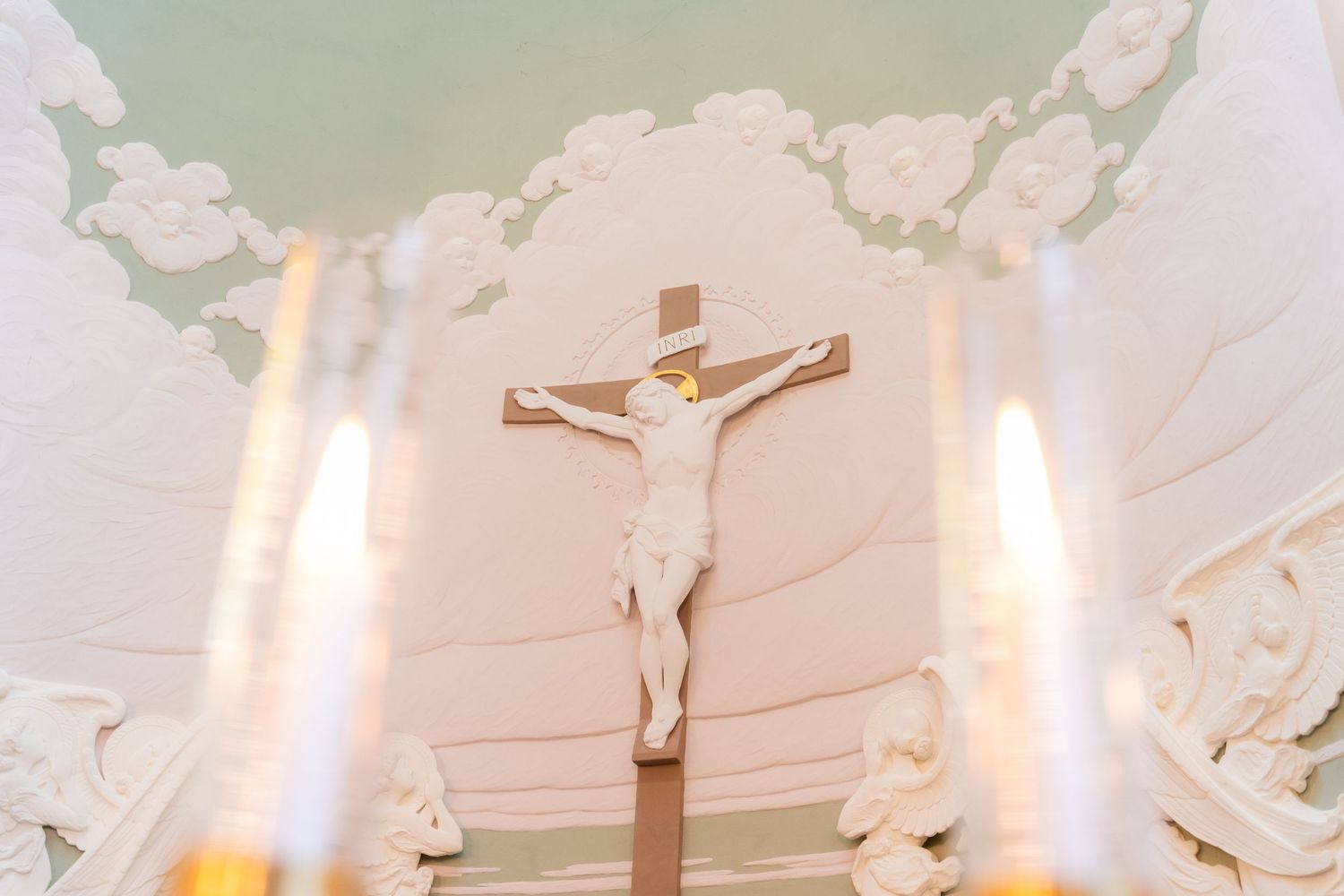 White crucifix sculpture on ornate church altar with glowing candles in foreground and decorative ceiling.
