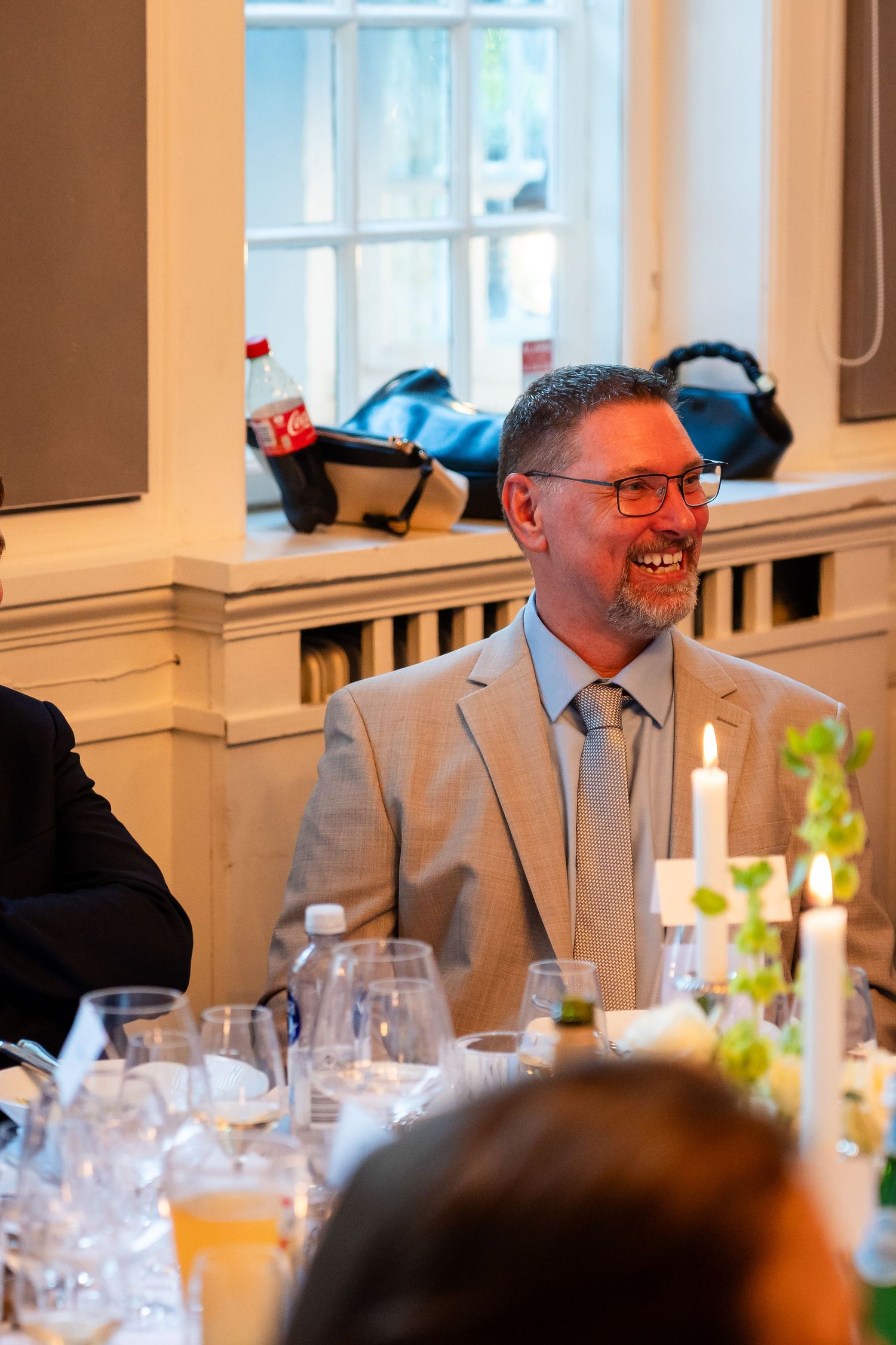 Smiling man with glasses and beard wearing a light gray suit at a formal dinner table with candles and glassware.