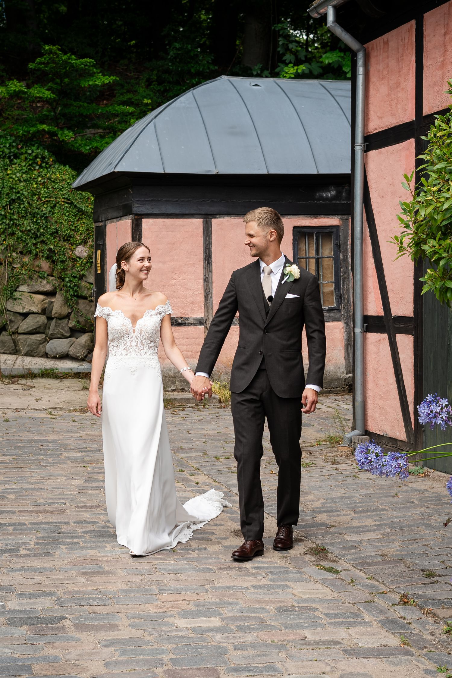 Bride and groom holding hands walking on cobblestone path beside pink timber-framed building on sunny wedding day.