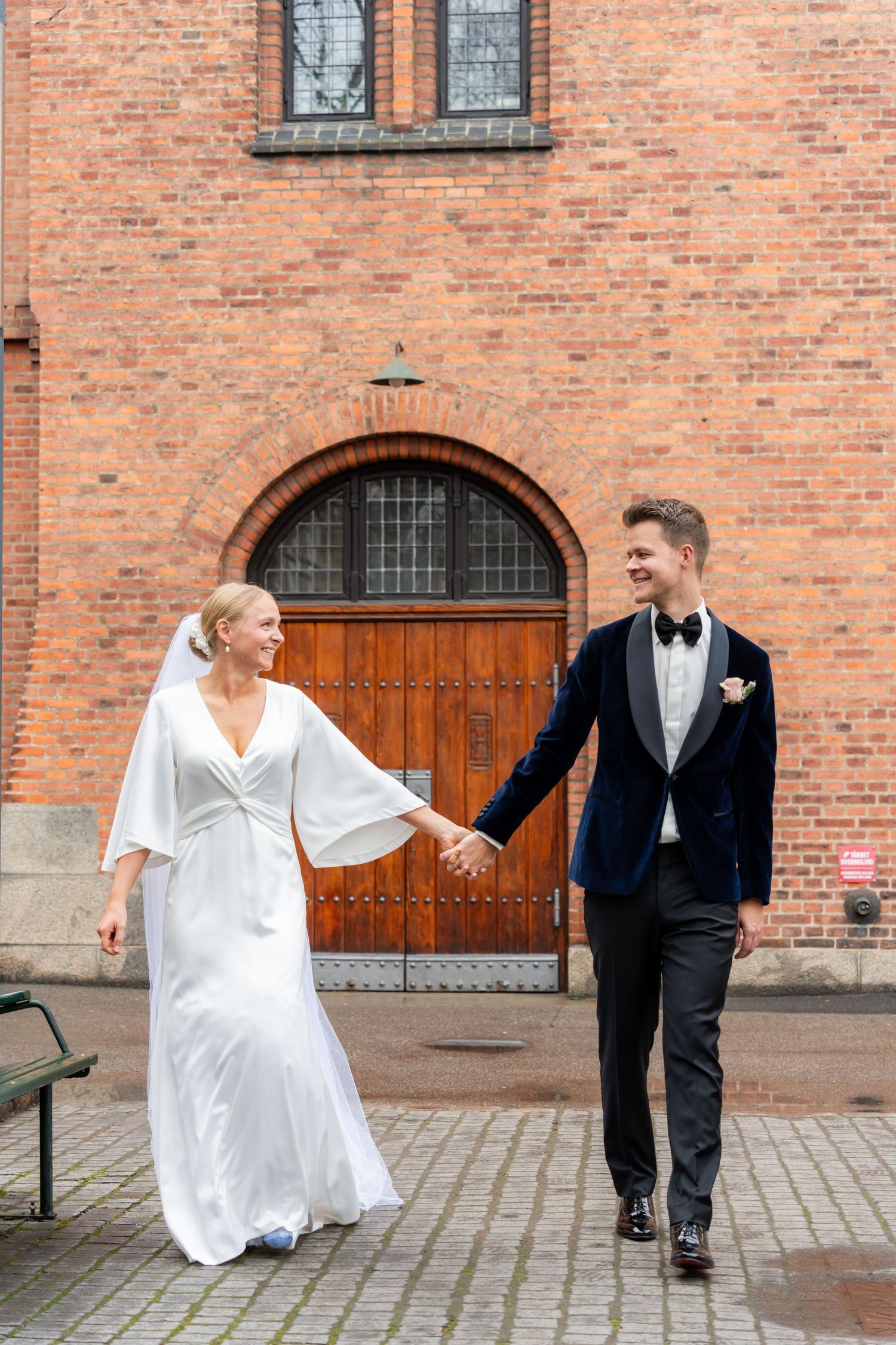 Bride and groom holding hands walking in front of a brick church with arched wooden doors on their wedding day.