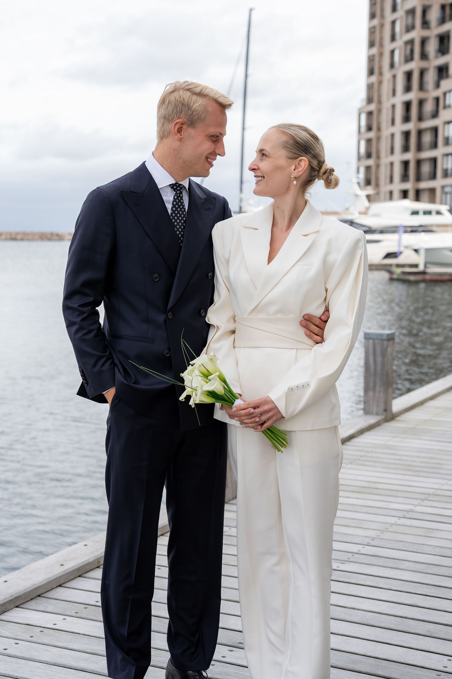 Elegant couple in wedding attire posing on a marina dock, groom in navy suit, bride in white pantsuit holding flowers.