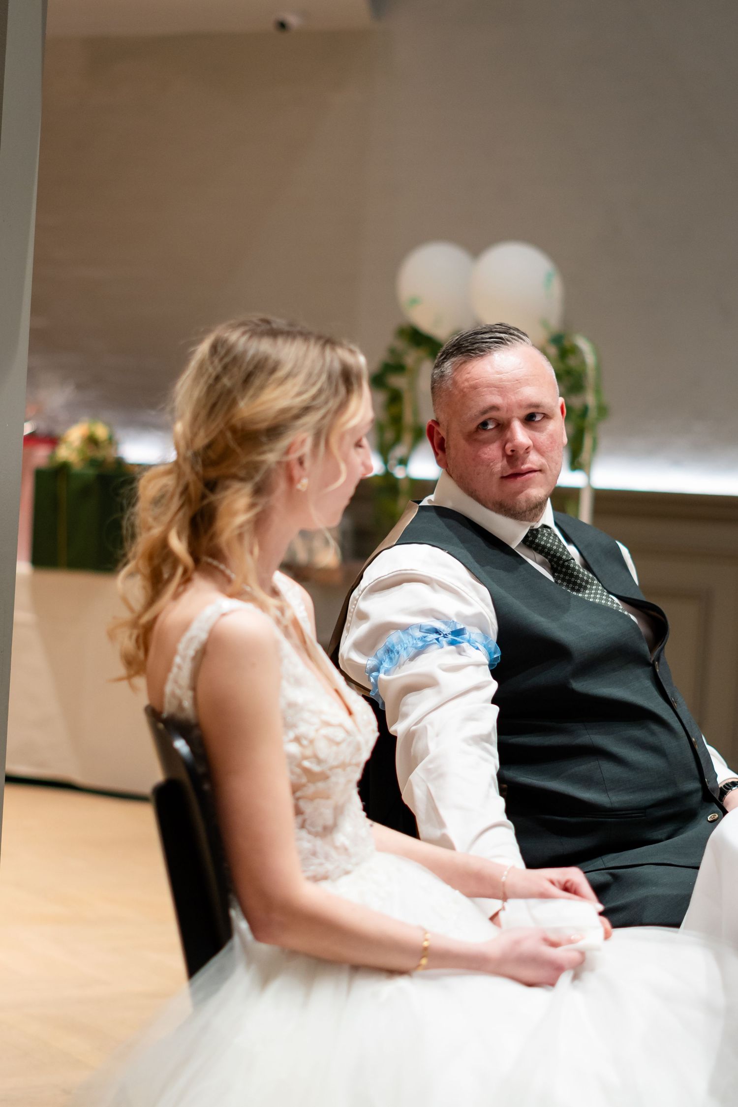 Bride and groom sitting together at wedding reception, groom gazing lovingly at blonde bride in white lace gown.