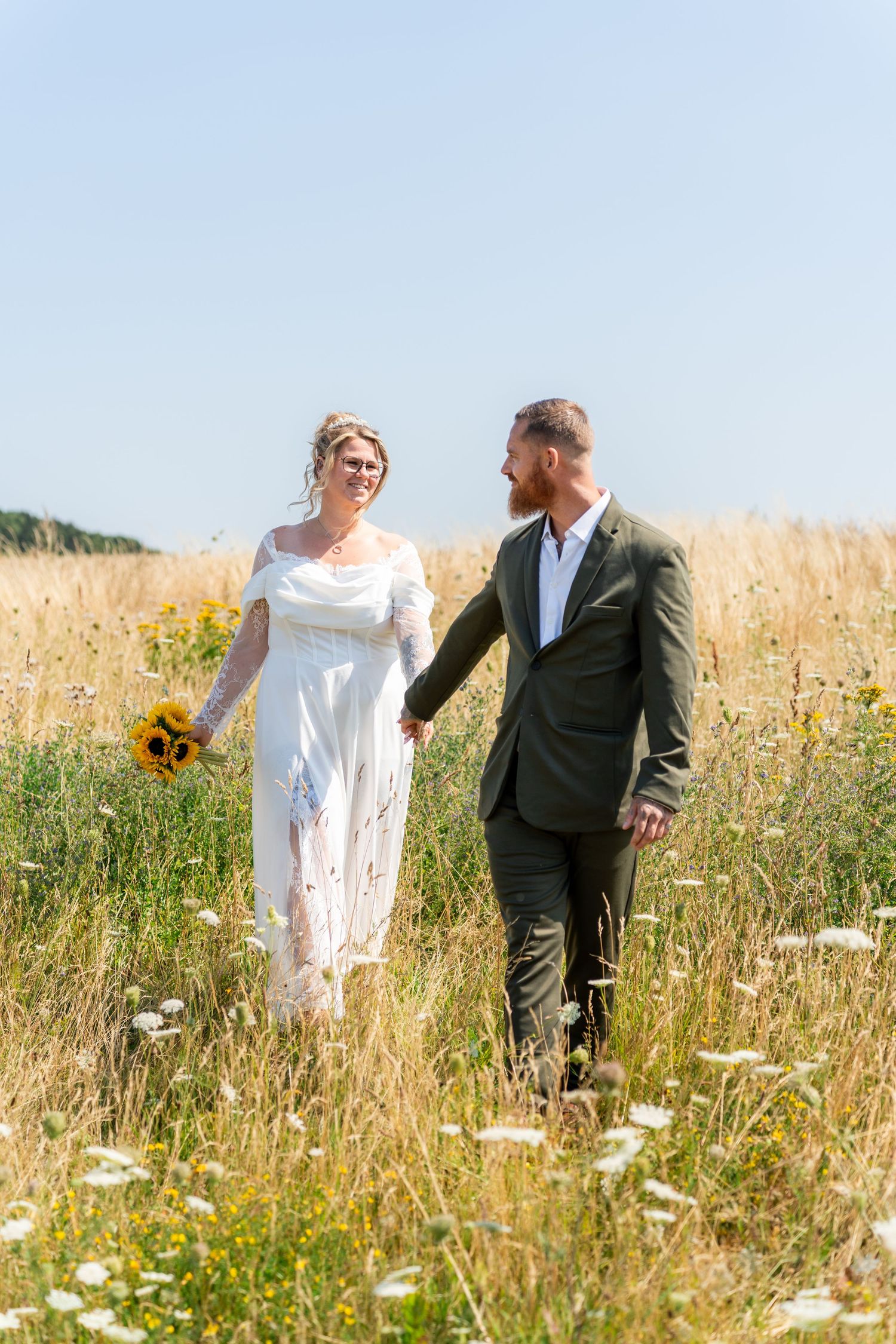 Bride and groom walking hand in hand through a sunny wildflower field, bride holding sunflower bouquet in white lace dress.