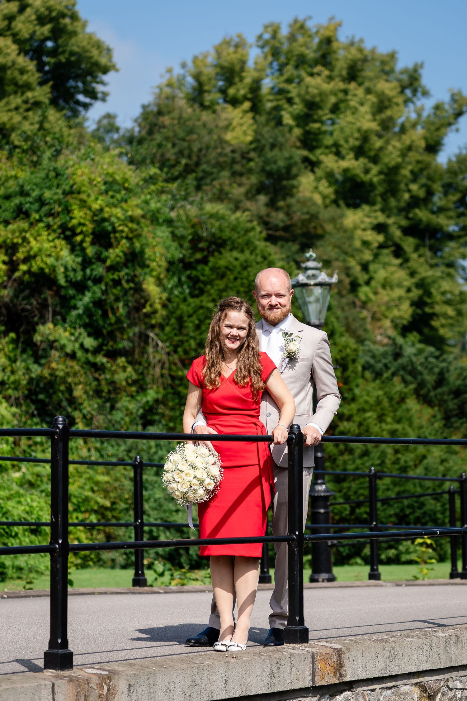 Bride in red dress holding bouquet stands with groom in gray suit on a bridge in a lush green park.