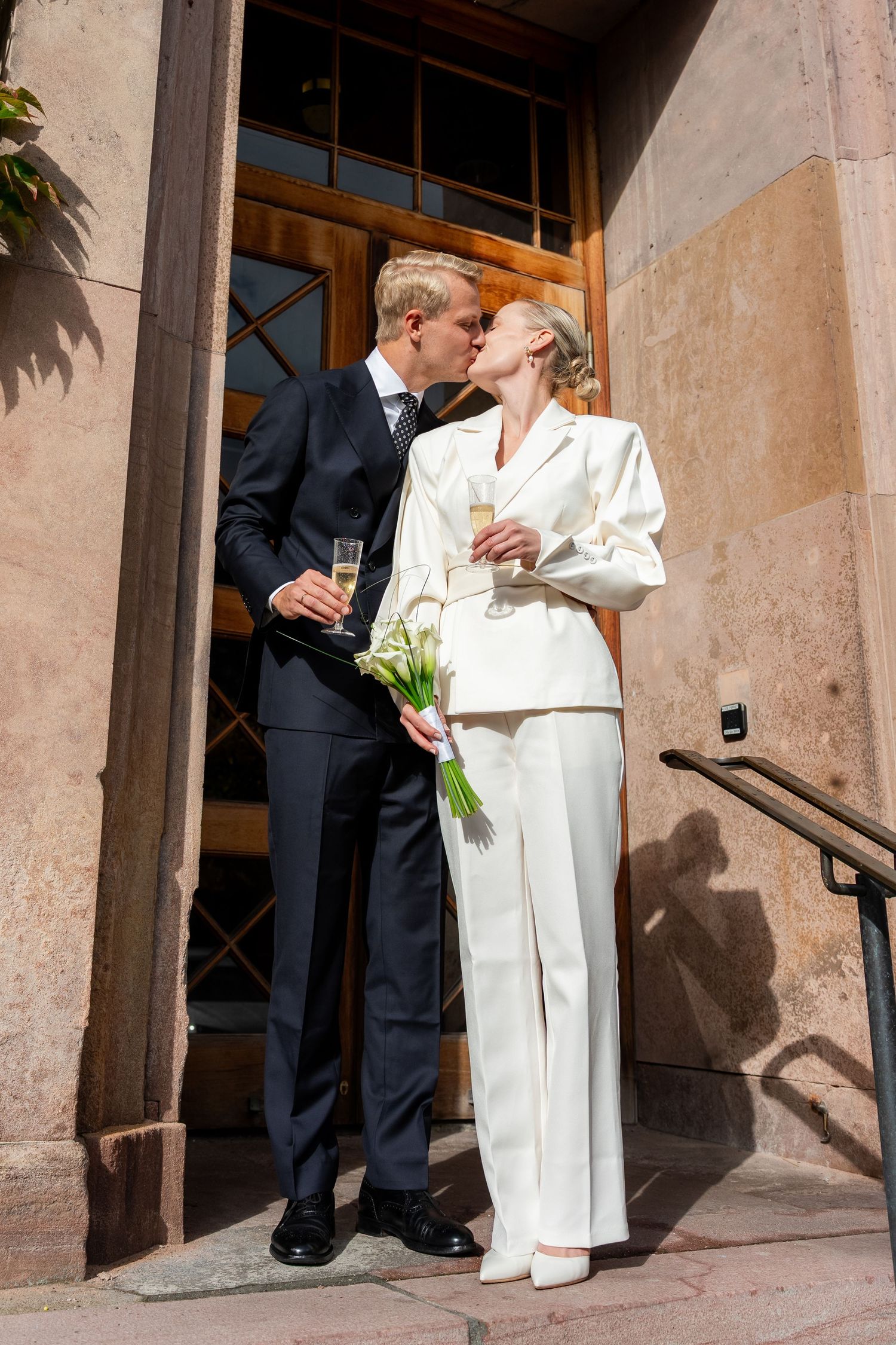 Newlywed couple kissing on building steps, bride in white suit holding calla lilies, groom in navy suit with champagne.