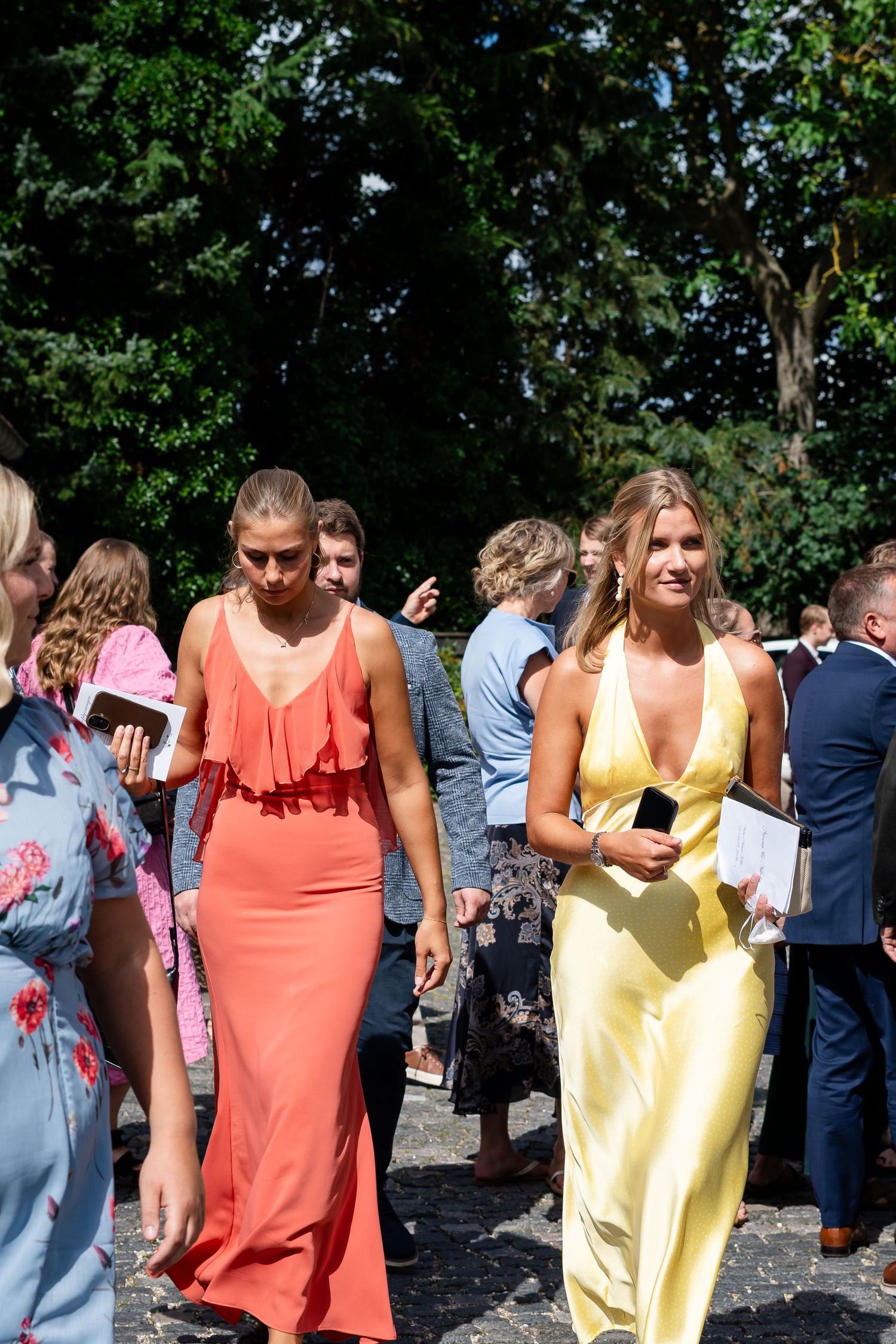 Two women in elegant maxi dresses, one coral and one yellow, walk among guests at an outdoor summer wedding.