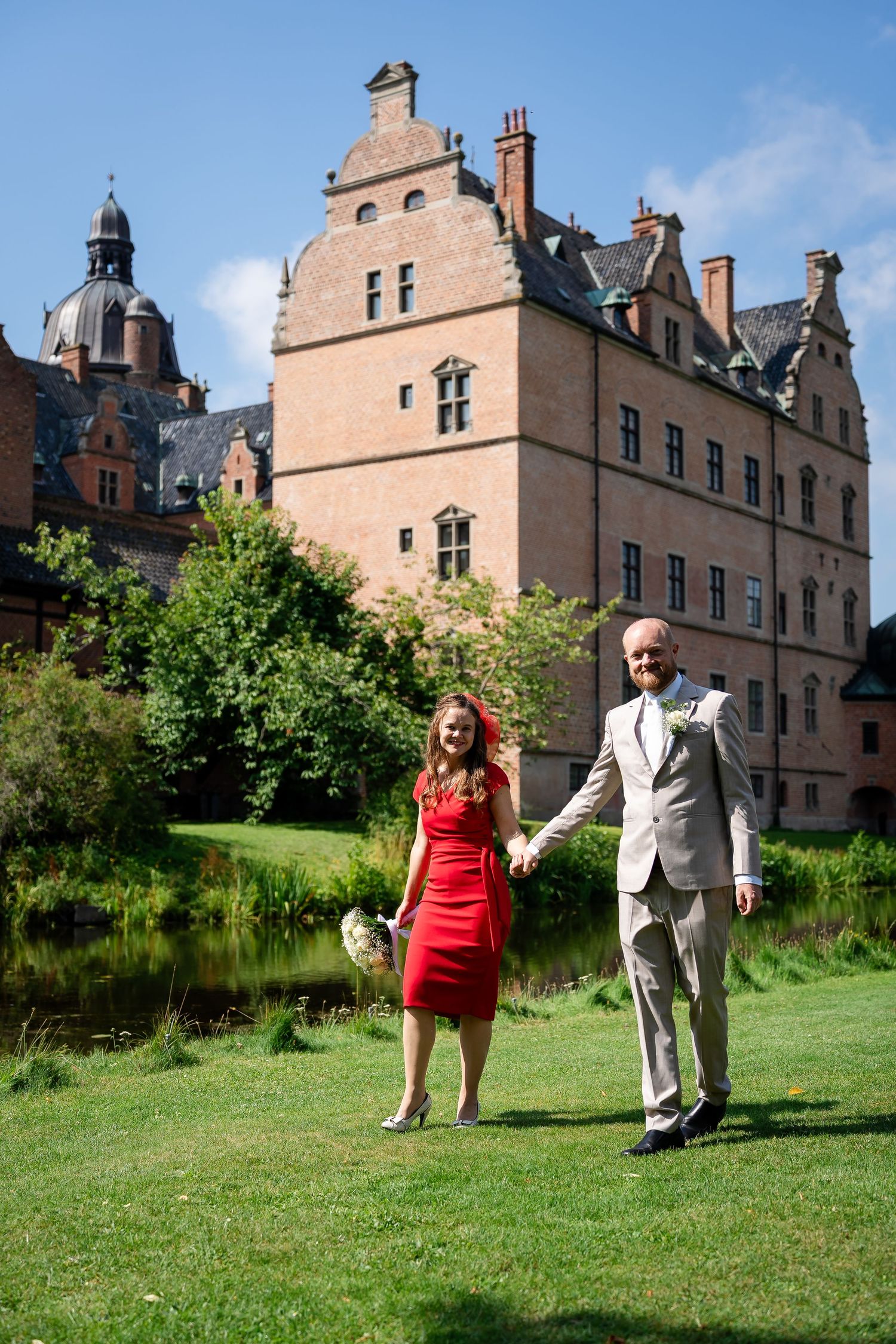 Couple in red dress and gray suit holding hands laughing on green lawn before historic brick castle on sunny day.