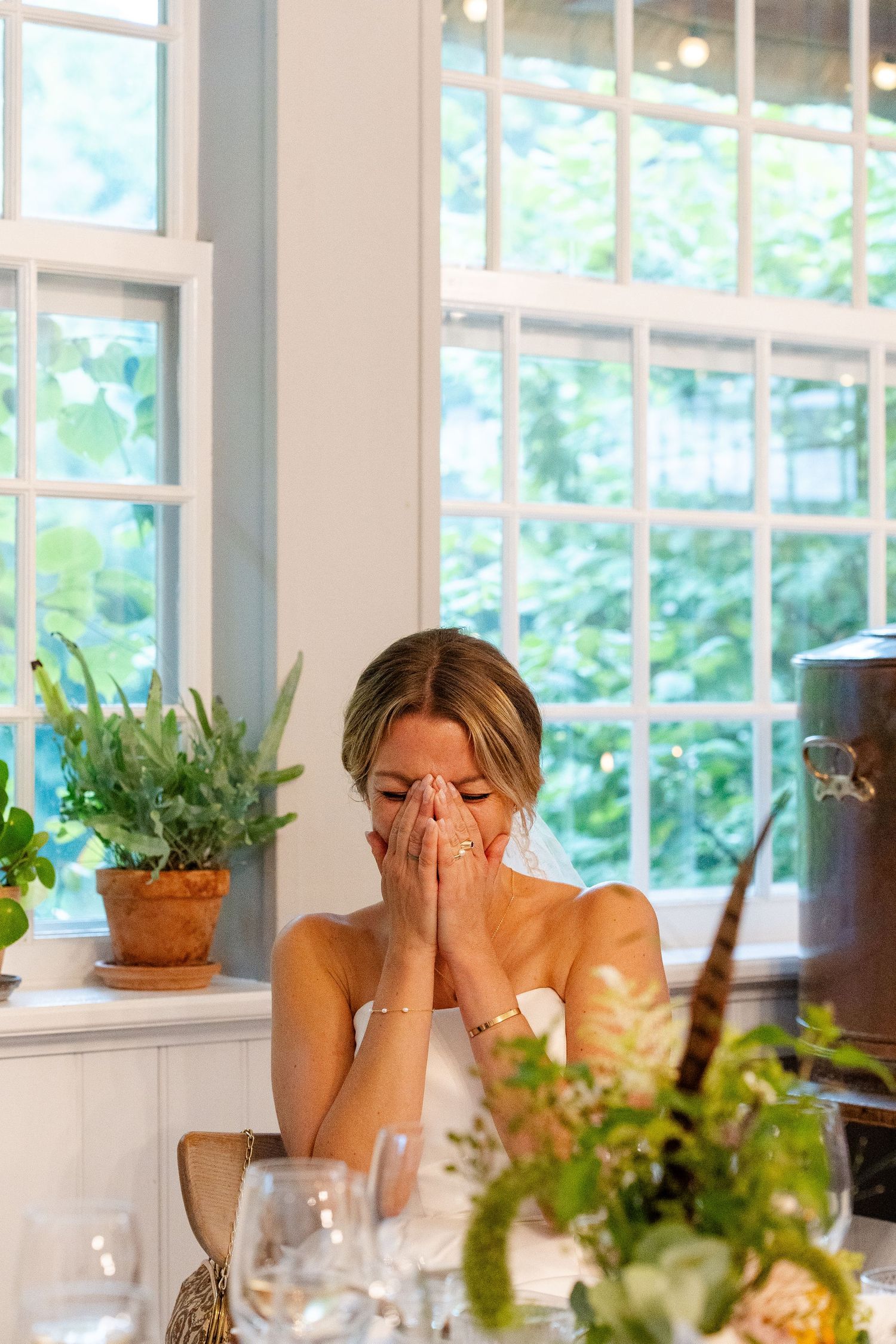 Emotional bride in strapless gown covers mouth with clasped hands at wedding reception table near sunlit windows.