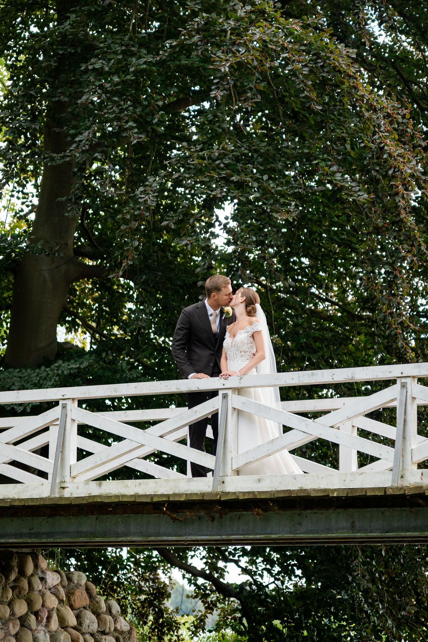 Bride and groom share a romantic moment on a white wooden bridge surrounded by lush green trees.