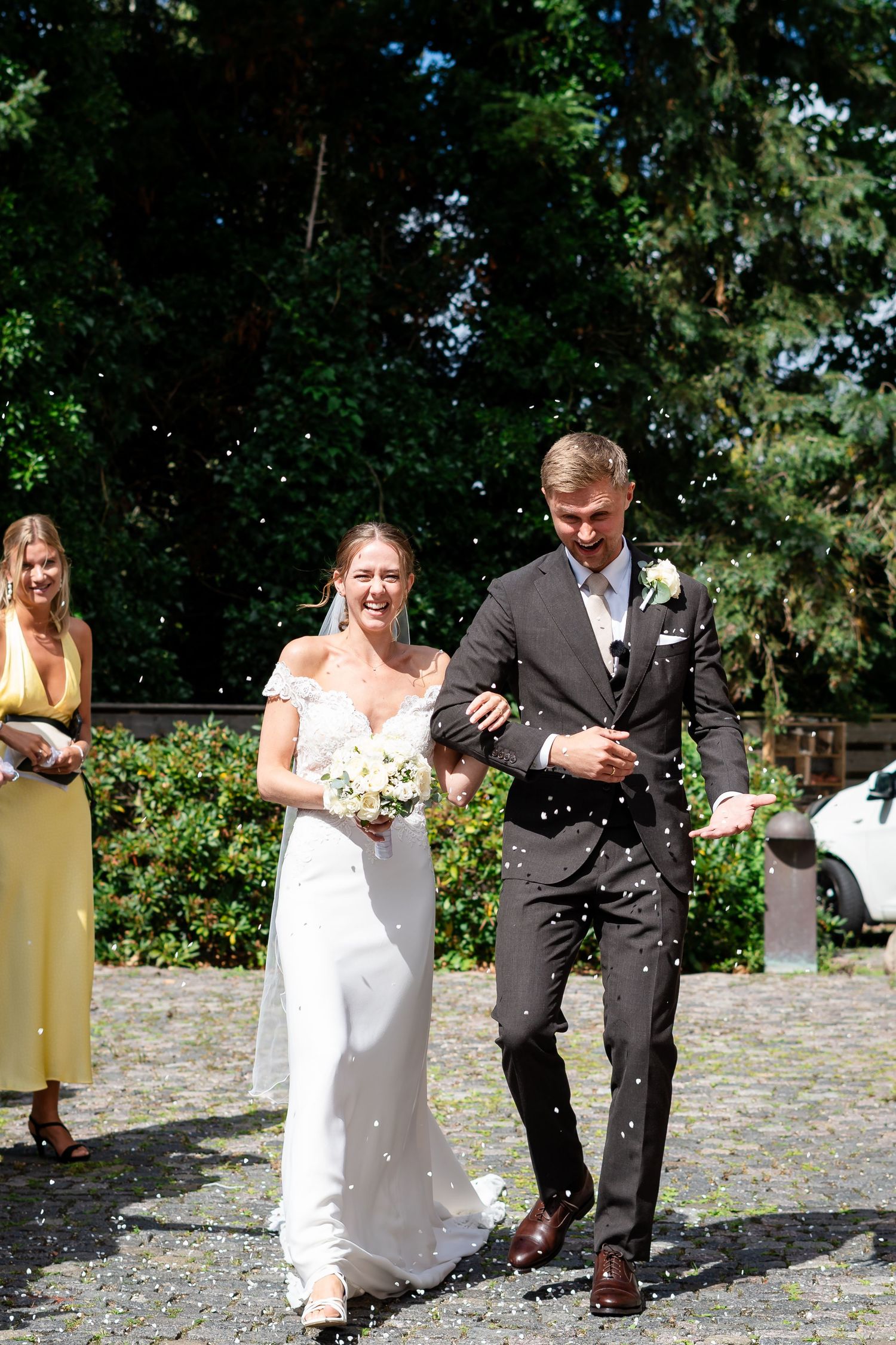 Bride and groom walk arm in arm through confetti shower outdoors, bride holding white bouquet in elegant white gown.