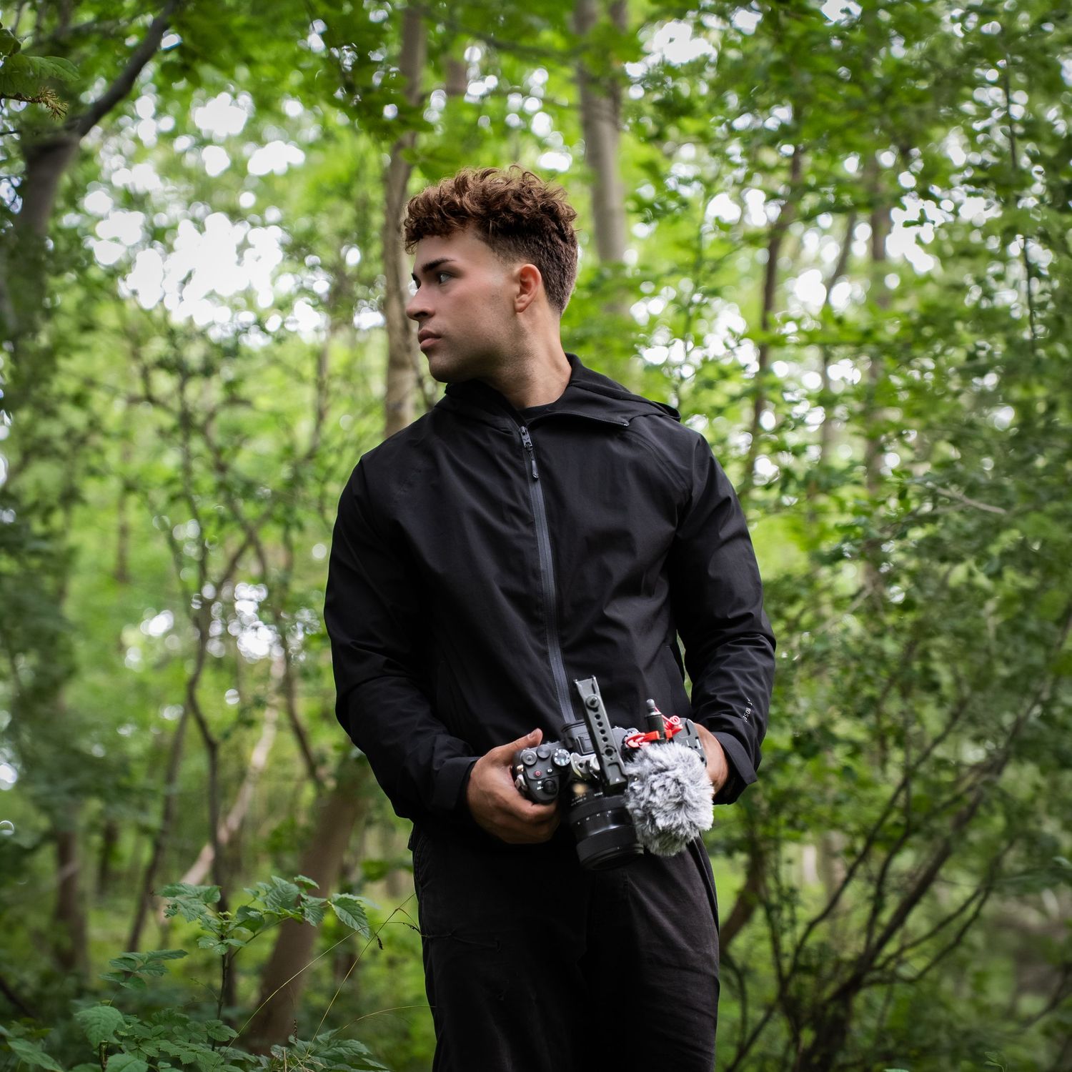 Young man in black jacket holding a camera in a lush green forest setting.