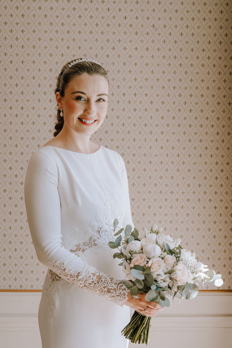 Bride in elegant long-sleeve white wedding gown holding delicate floral bouquet.