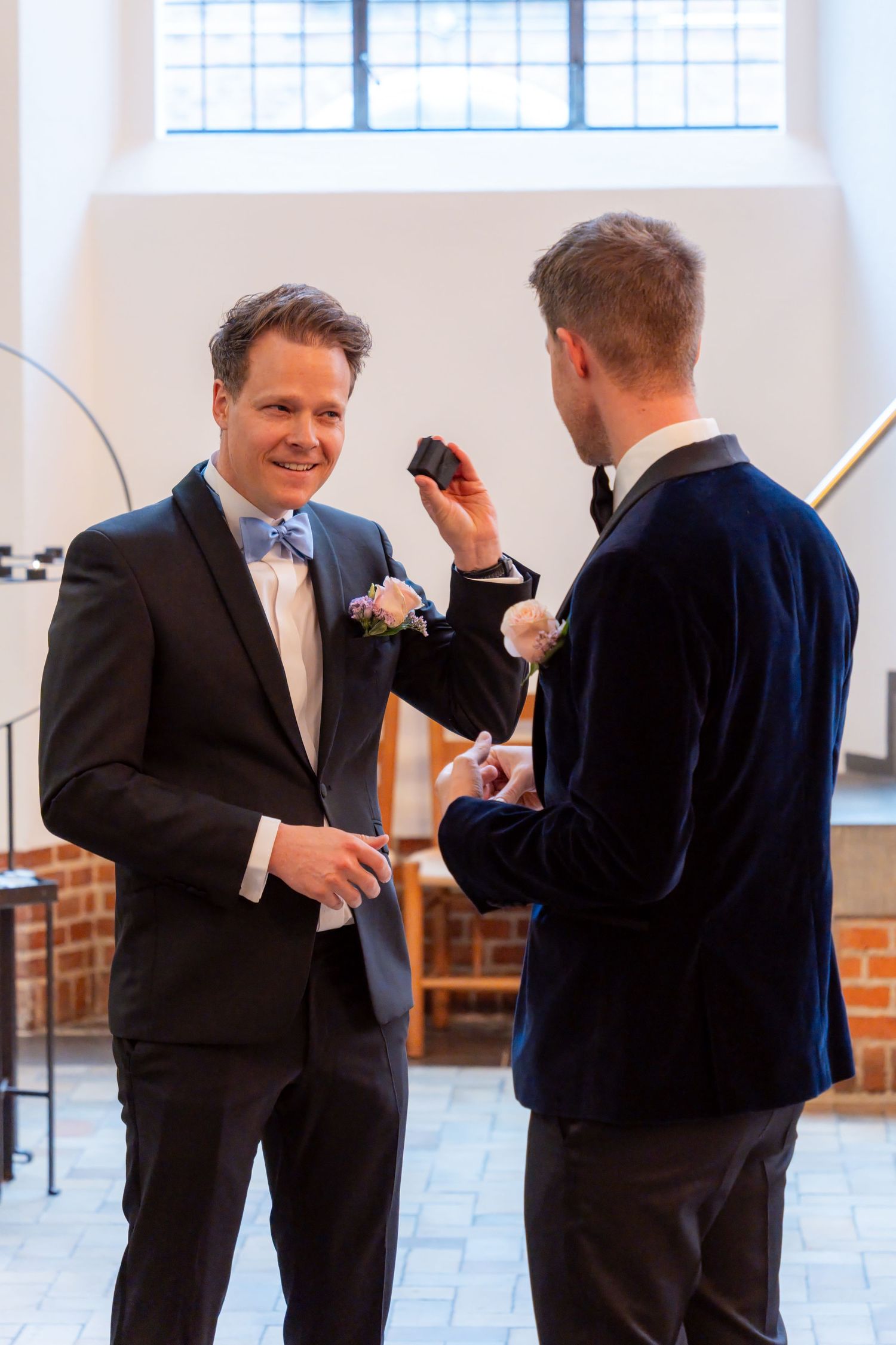 Two grooms exchanging rings during a wedding ceremony, wearing suits with boutonnieres in a church.