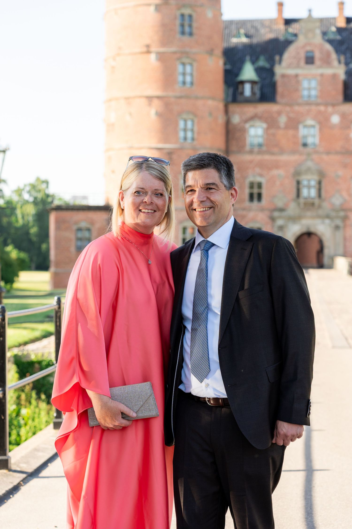 Smiling couple in formal attire posing in front of a historic red brick castle on a sunny day.