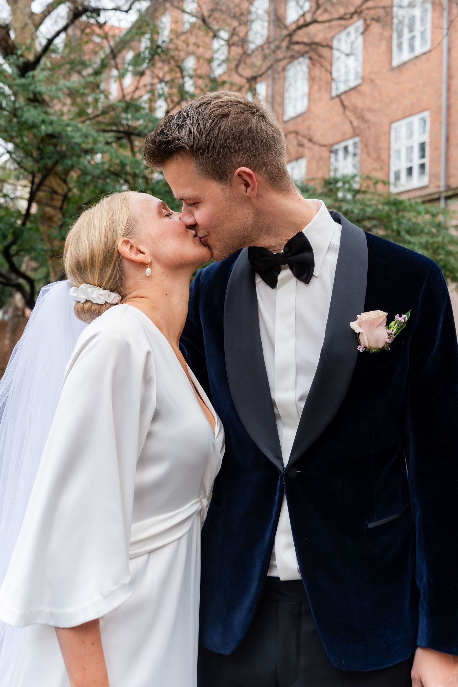 Bride and groom sharing a kiss outdoors, wearing white wedding dress with veil and navy blue velvet tuxedo with bow tie.