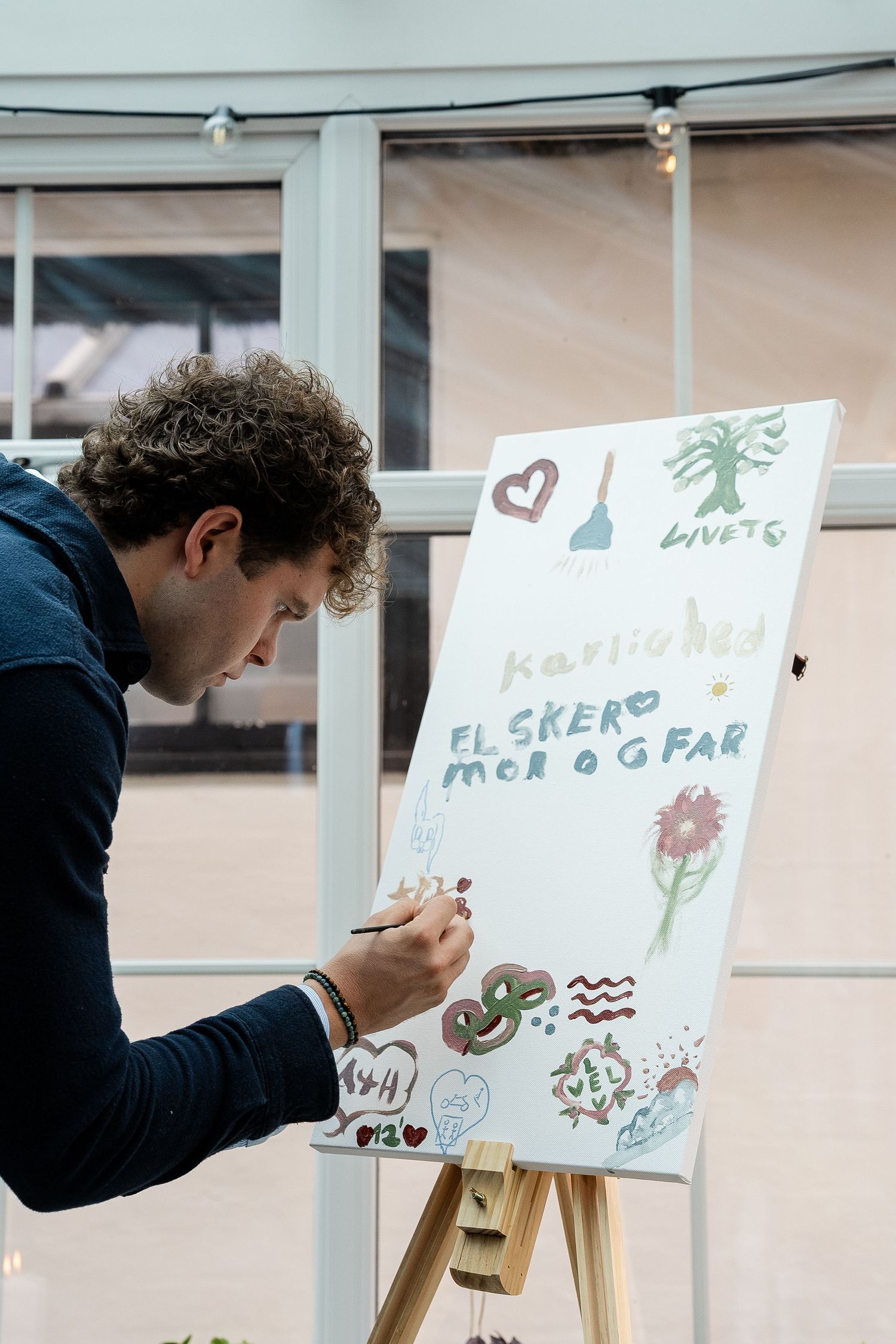 Man writing on decorated easel canvas with colorful drawings and Danish text at an indoor event.