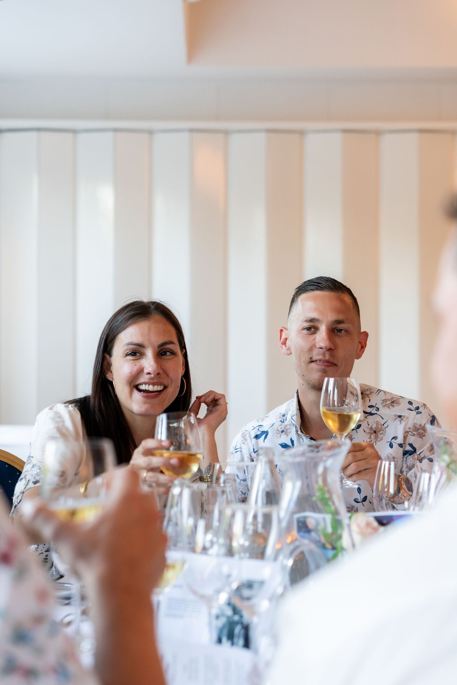 A smiling couple holding white wine glasses at an elegant restaurant dining table during a social gathering.