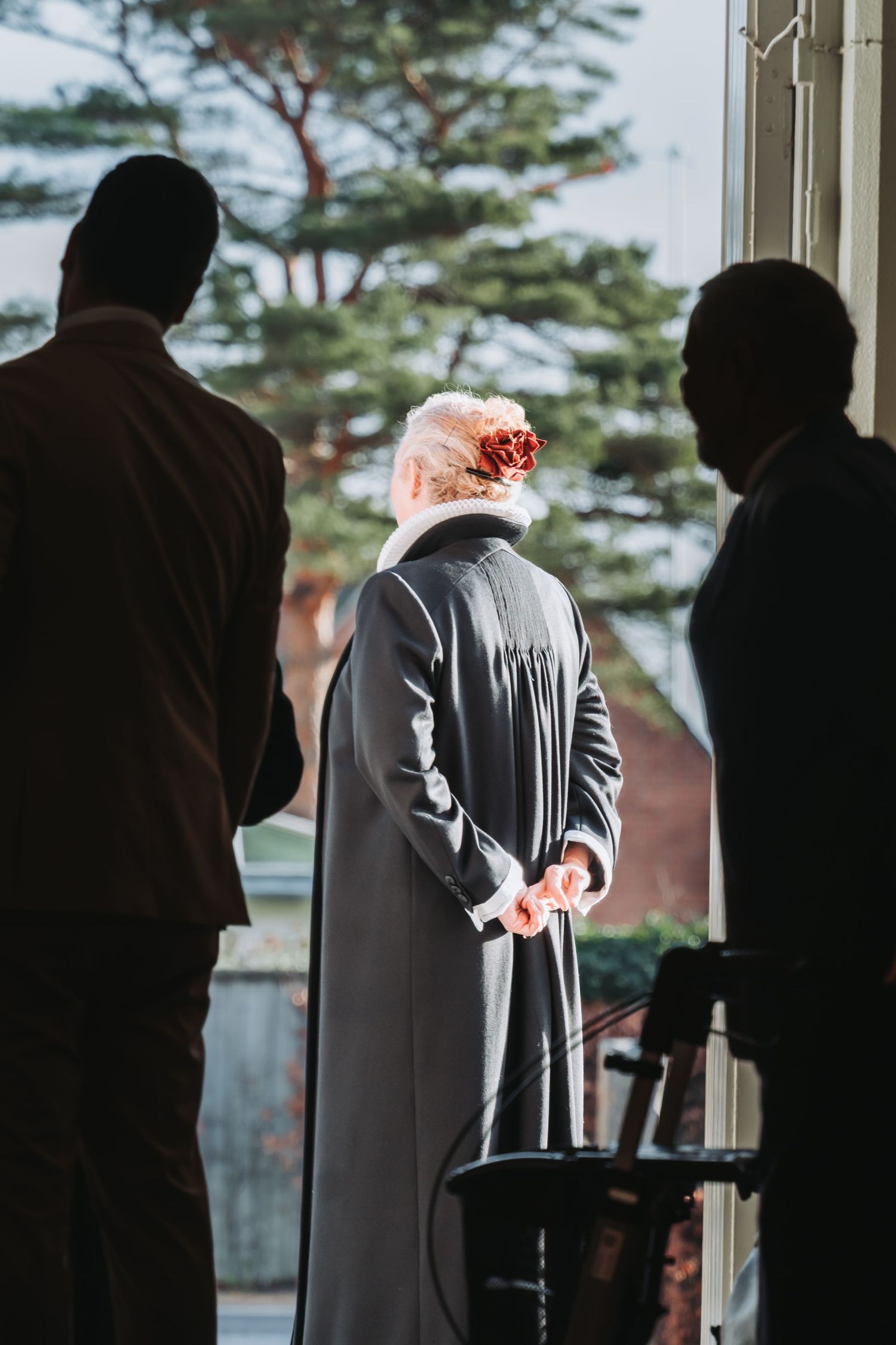 Elderly woman in gray coat with floral hair accessory stands between two silhouetted figures near a bright window.