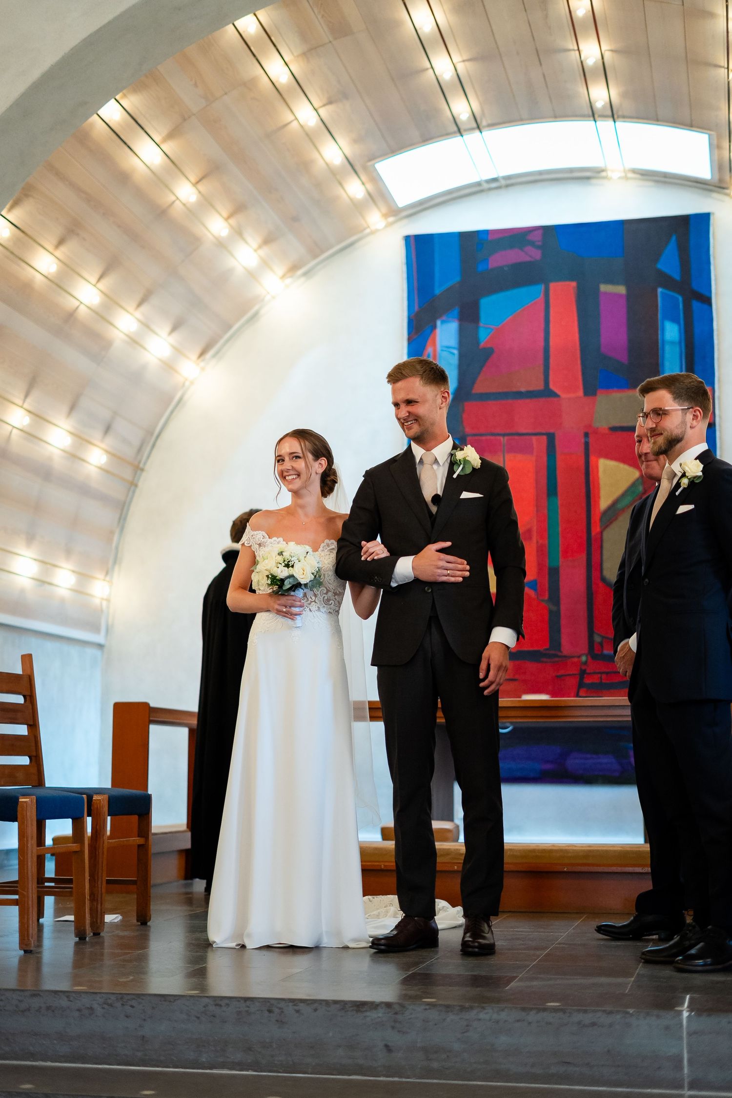 Bride and groom smiling at wedding ceremony in modern chapel with colorful stained glass window and arched ceiling.