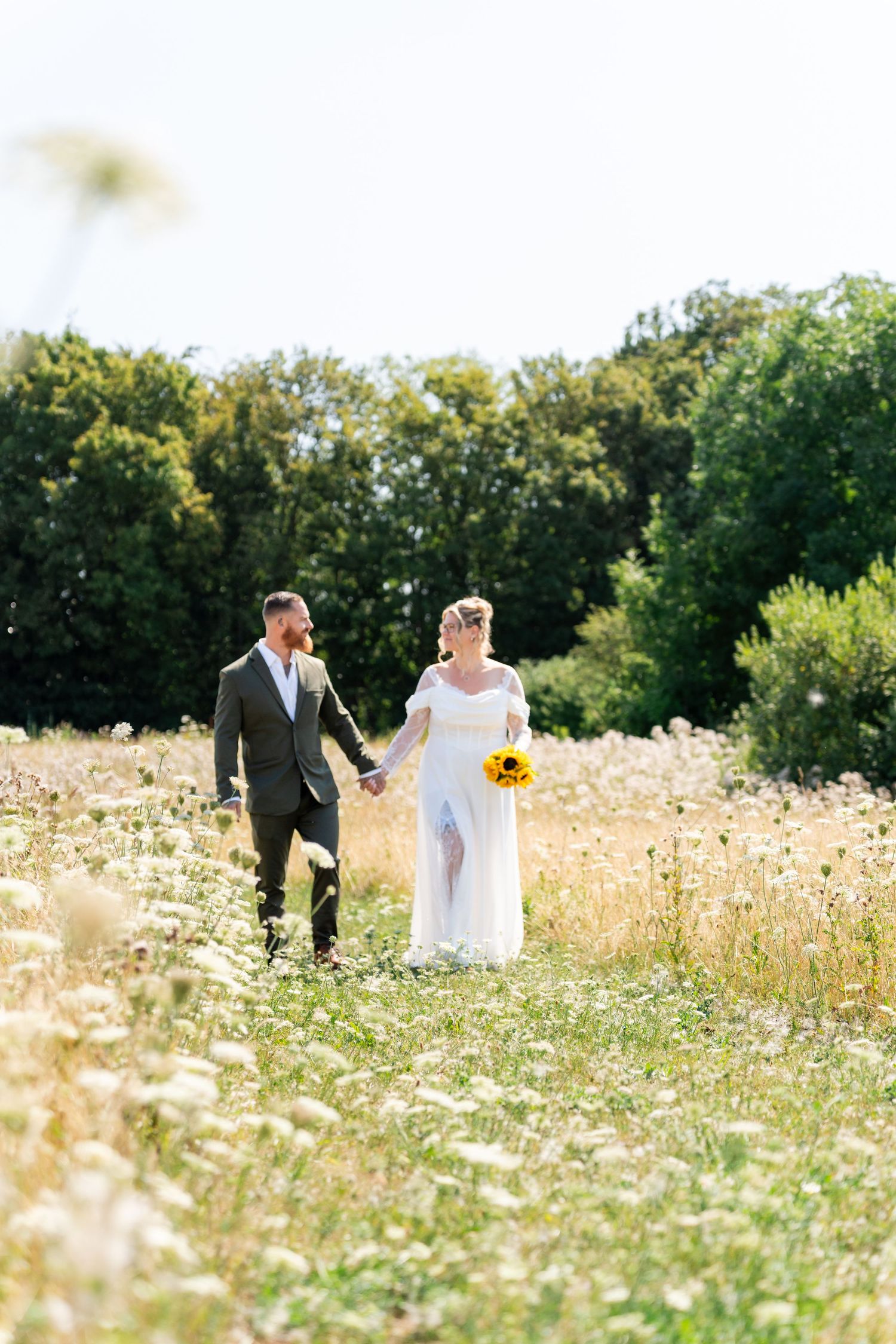 Bride and groom walk hand-in-hand through a sunlit wildflower meadow, bride holding a sunflower bouquet.