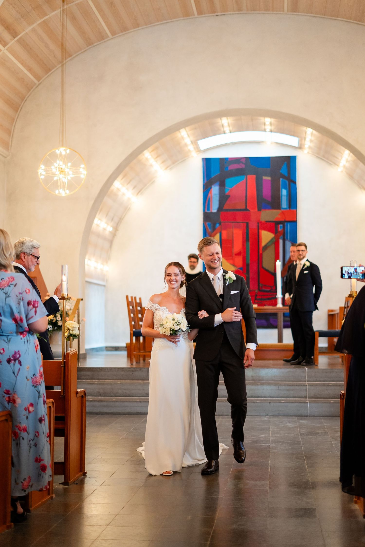 Bride and groom walk down church aisle smiling, colorful stained glass window behind them during wedding ceremony.