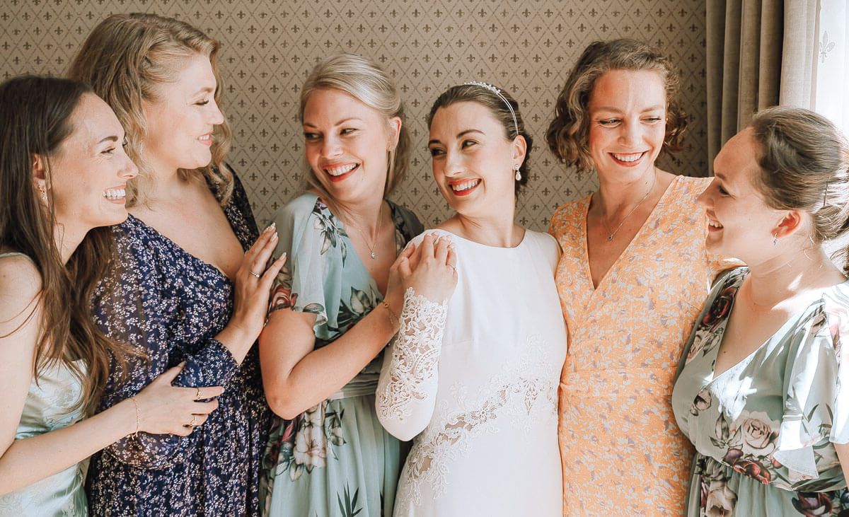 Row of bridesmaids in pastel dresses posing against patterned wallpaper.