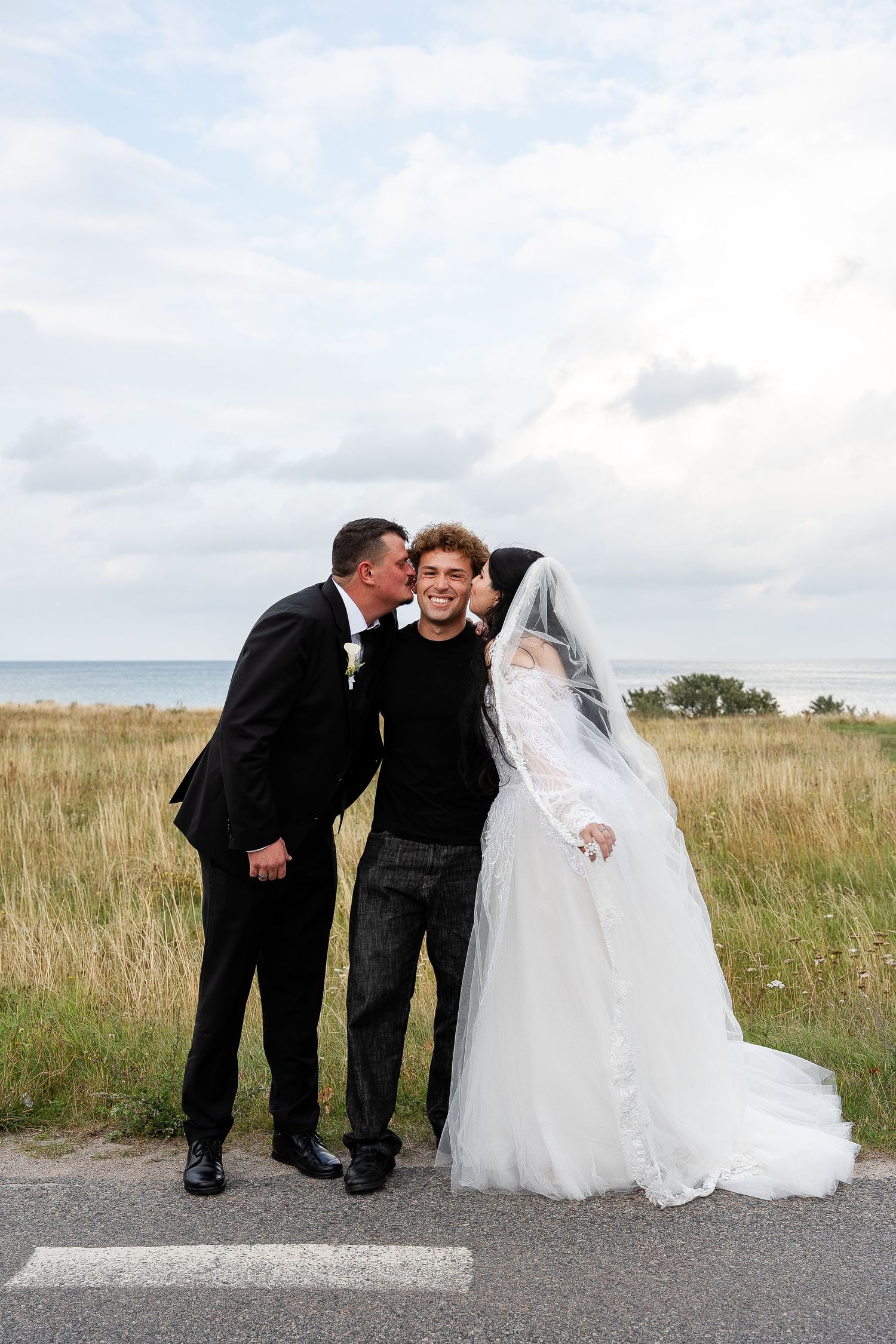 A bride and groom share a wedding day kiss outdoors near a coastal field under a cloudy sky.