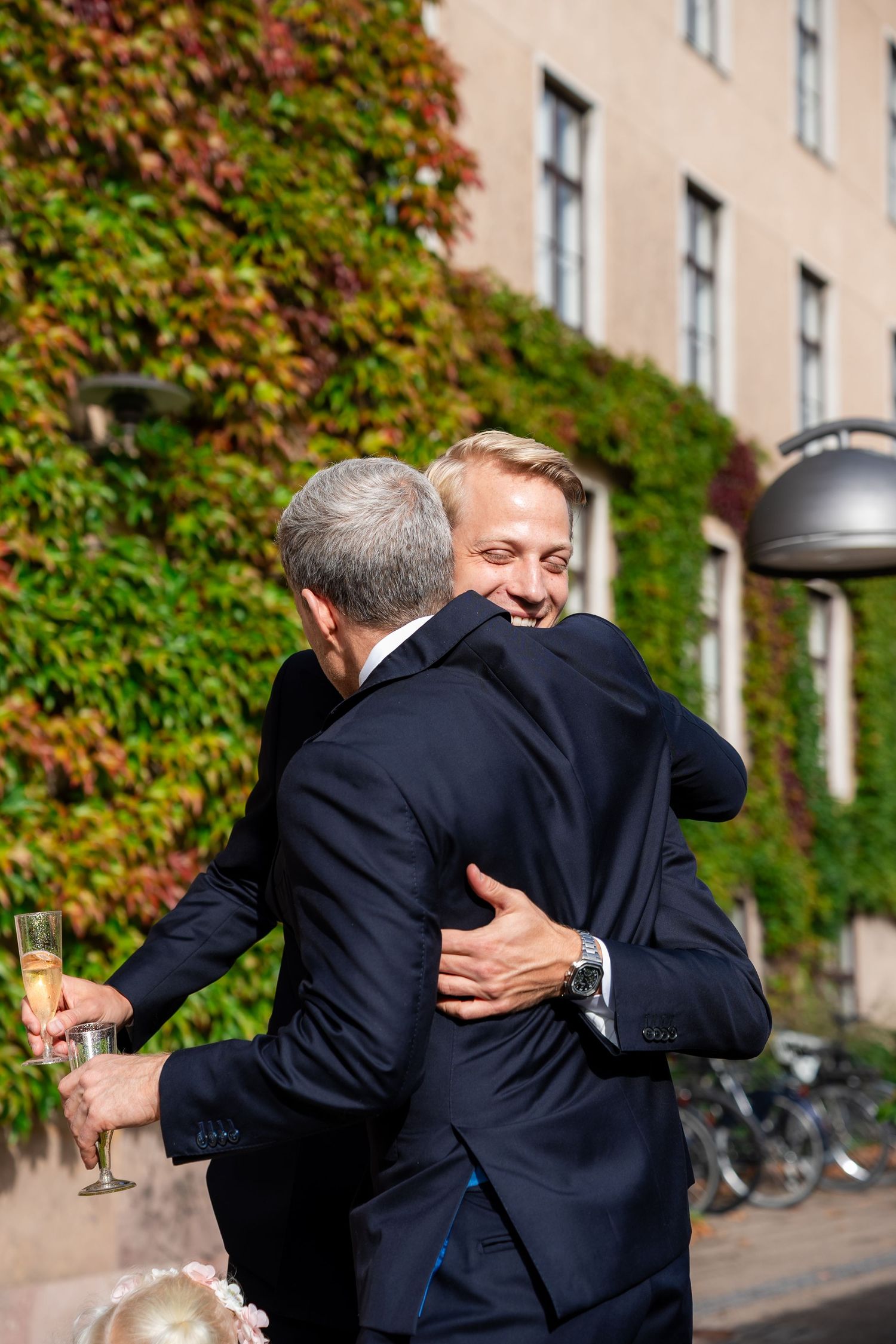 Two men in dark suits share a warm hug outdoors, one holding champagne, with ivy-covered building in background.