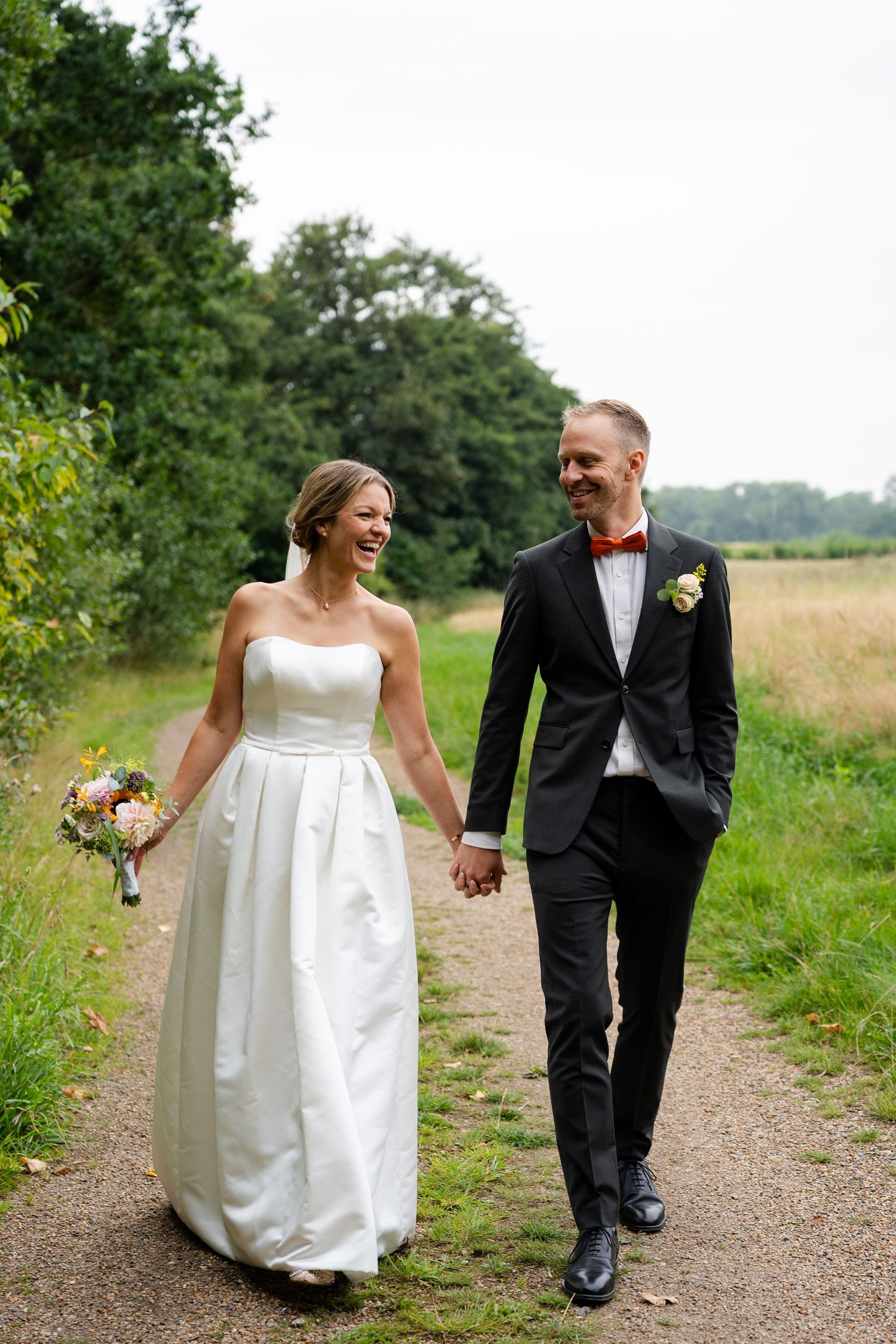 A laughing bride in a white strapless gown holds hands with a groom in a black suit and bow tie while walking outdoors.