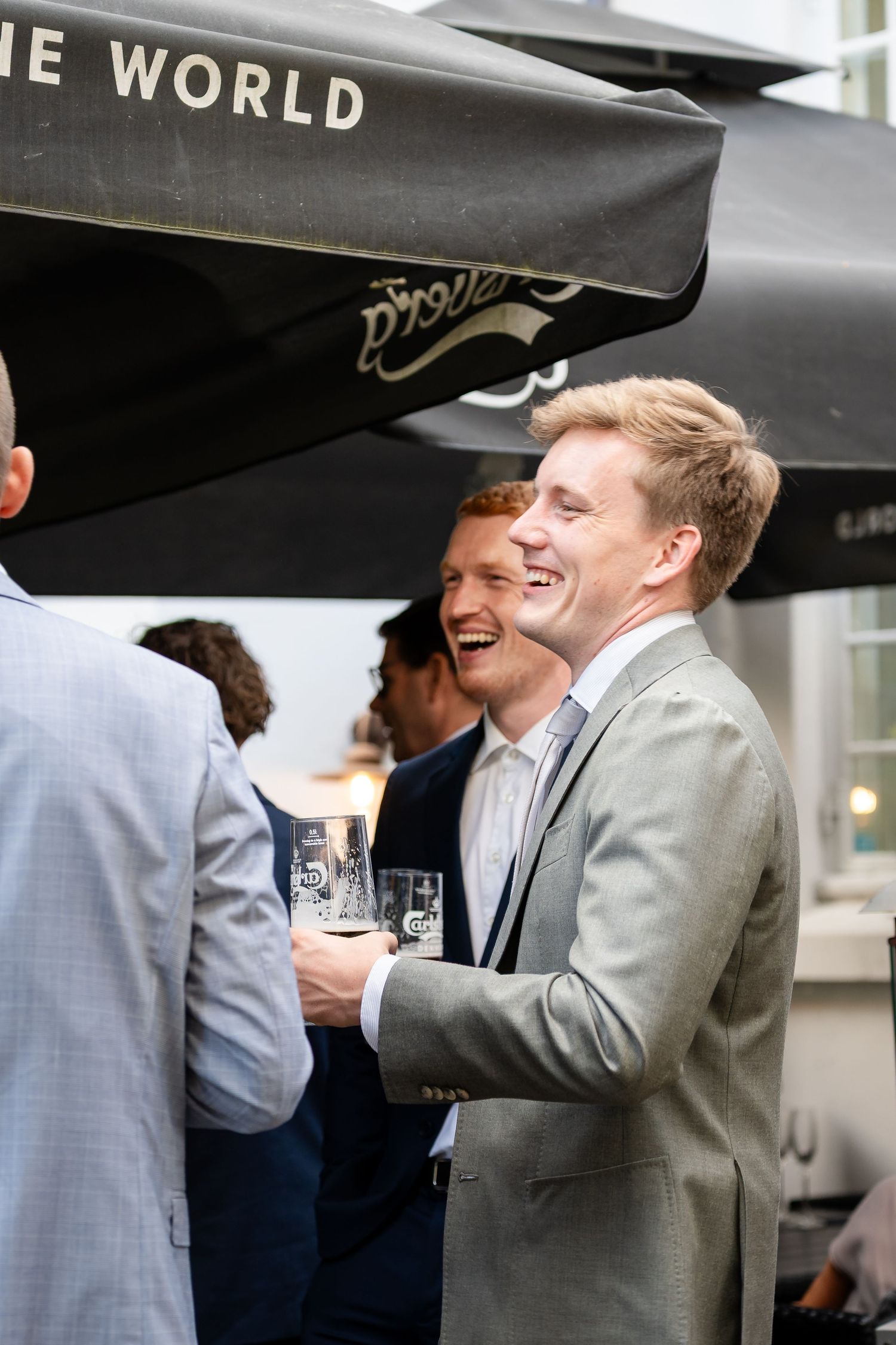 Two young men in suits laughing and holding drinks at an outdoor networking event under a black umbrella.