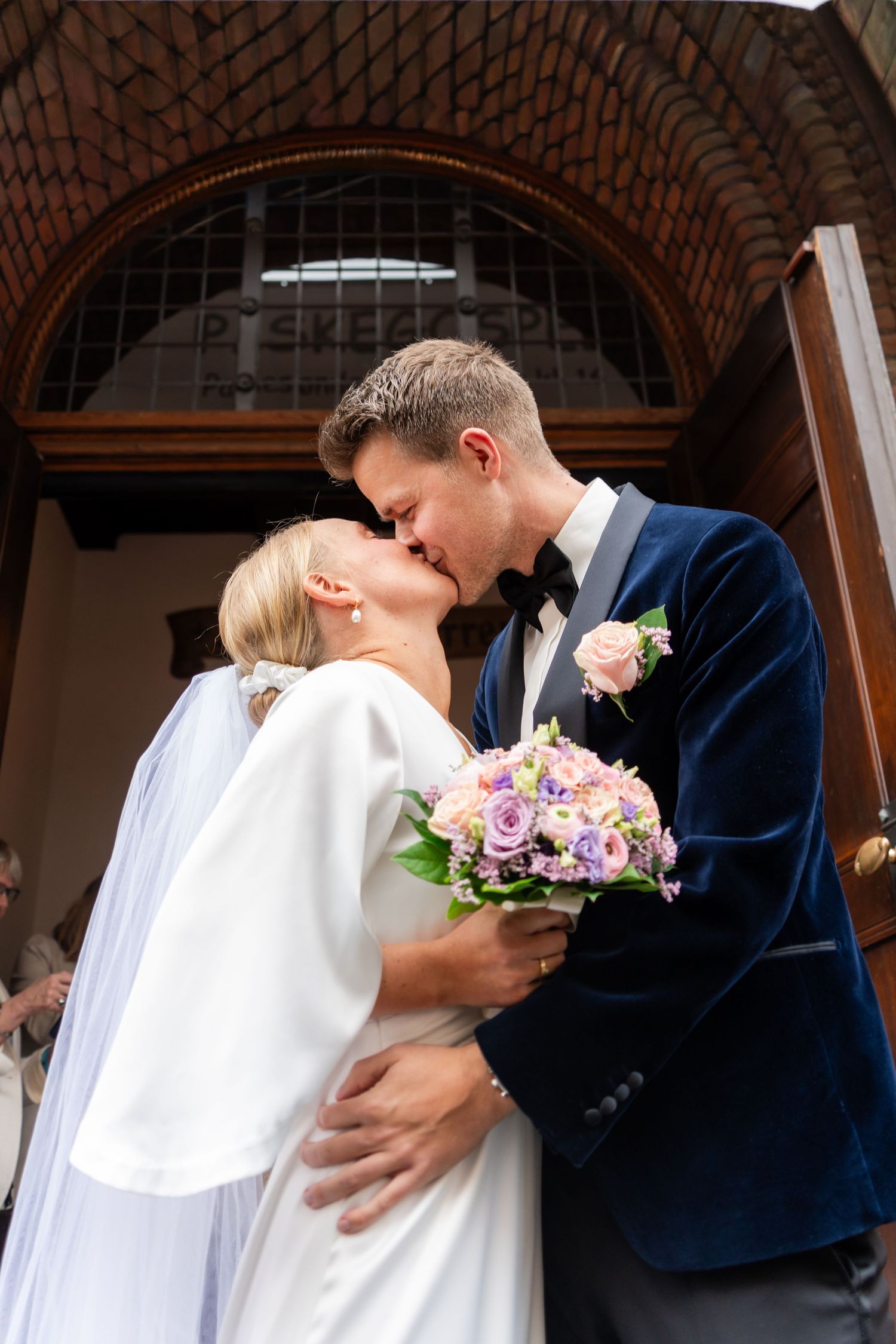 Bride and groom share a first kiss in wedding attire outside a church, bride holding a purple and pink bouquet.