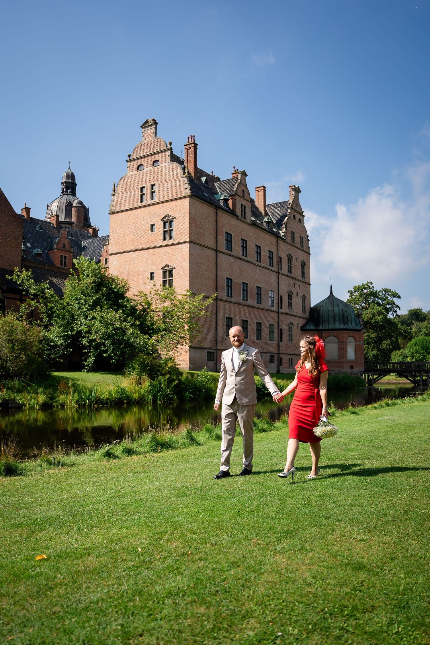 Couple holding hands walking on green lawn before a grand historic red brick castle on a sunny day.