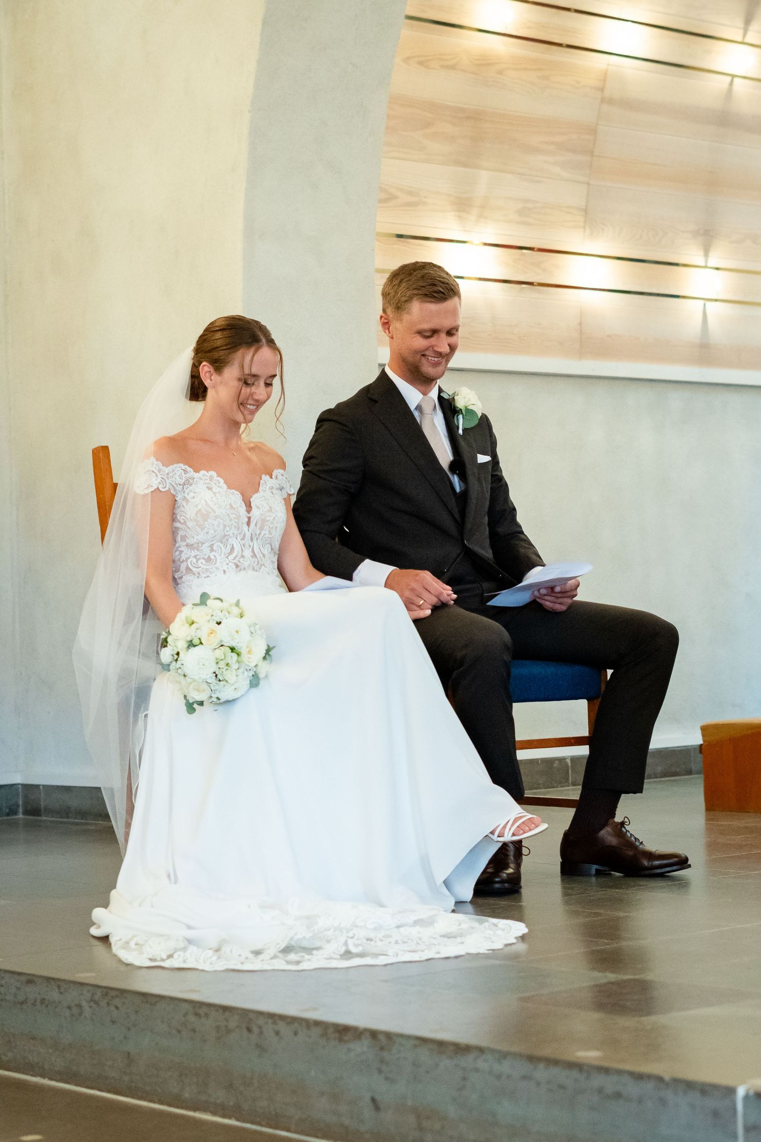 Smiling bride and groom sitting together during wedding ceremony, bride holding white bouquet in elegant lace gown with veil.