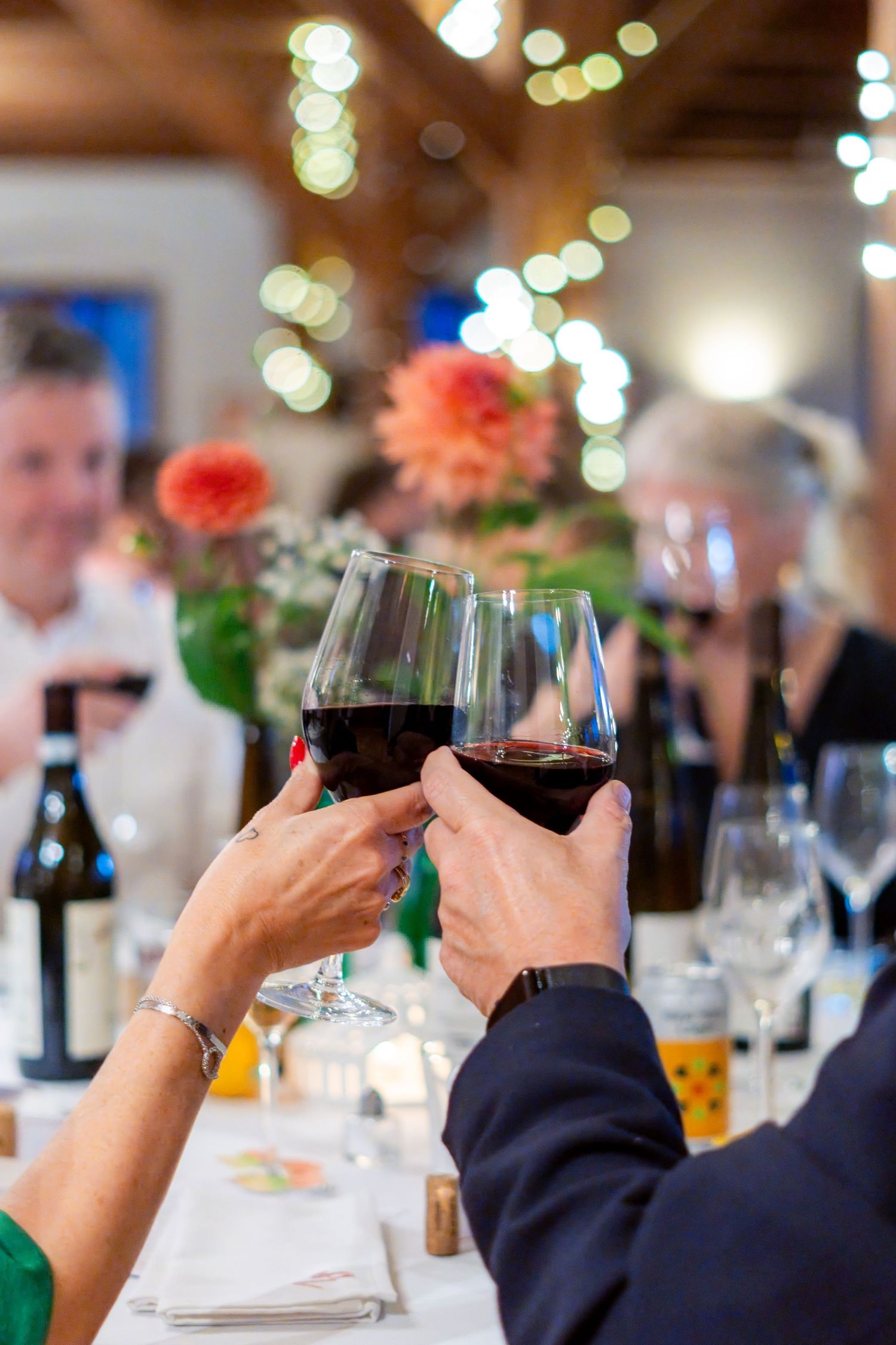 Two people clinking glasses of red wine at an elegant dinner party with bokeh lights in the background.