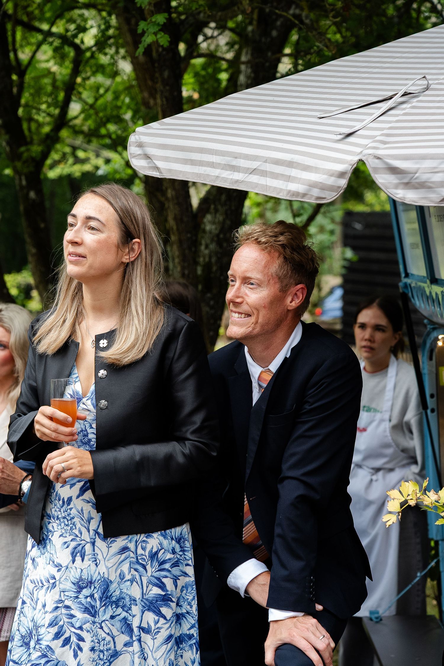 A smiling man and woman in elegant attire enjoy an outdoor garden party, with lush green trees in the background.