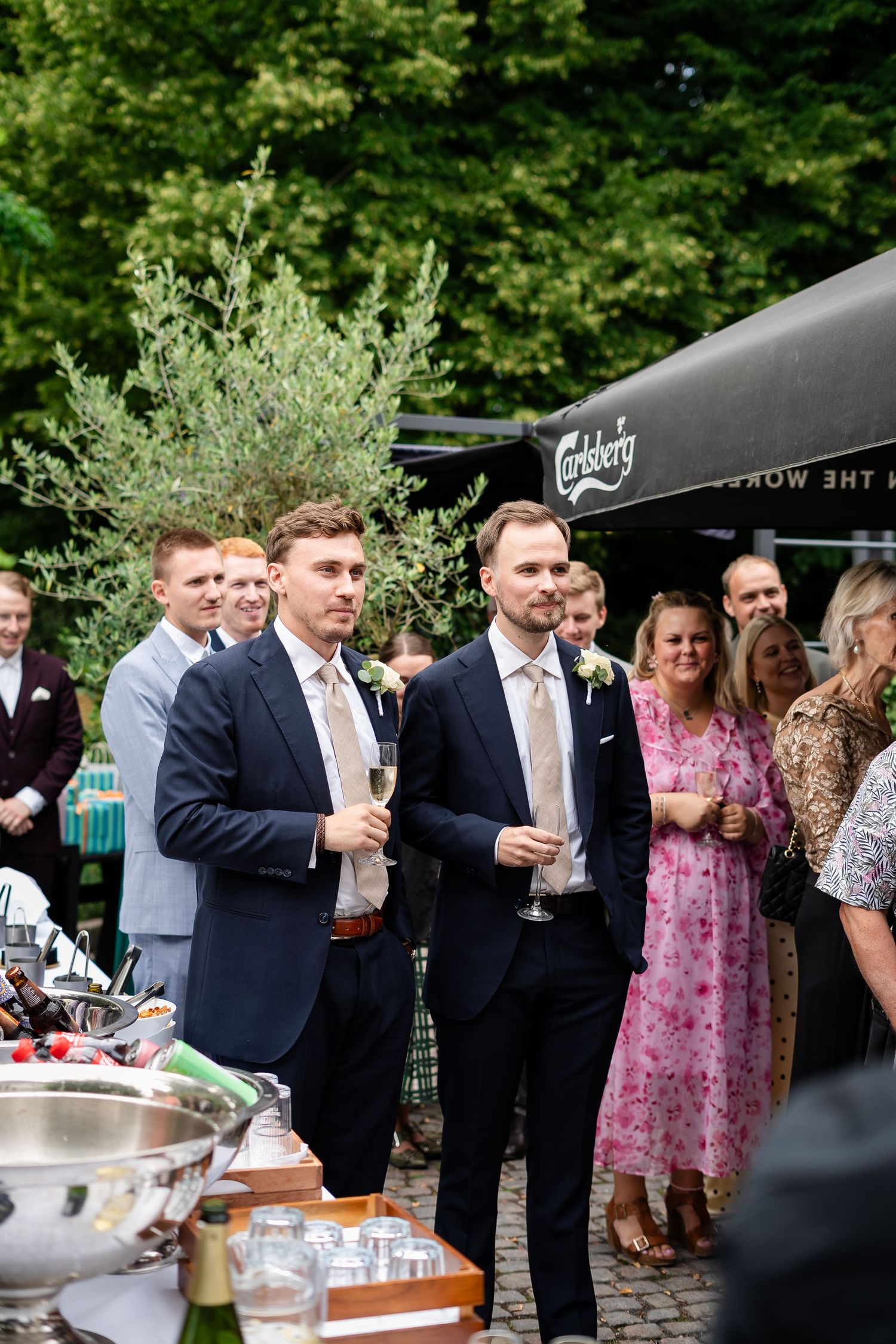 Groomsmen in navy suits holding champagne glasses at an outdoor wedding reception with guests mingling nearby.