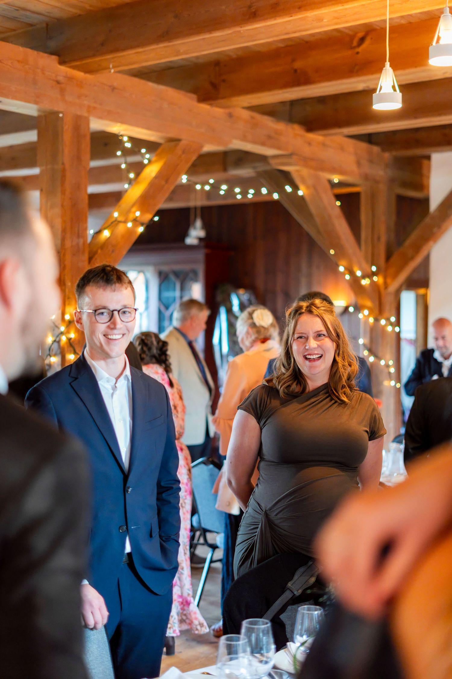 Smiling couple in elegant attire socializing at a warmly lit rustic venue decorated with fairy lights.