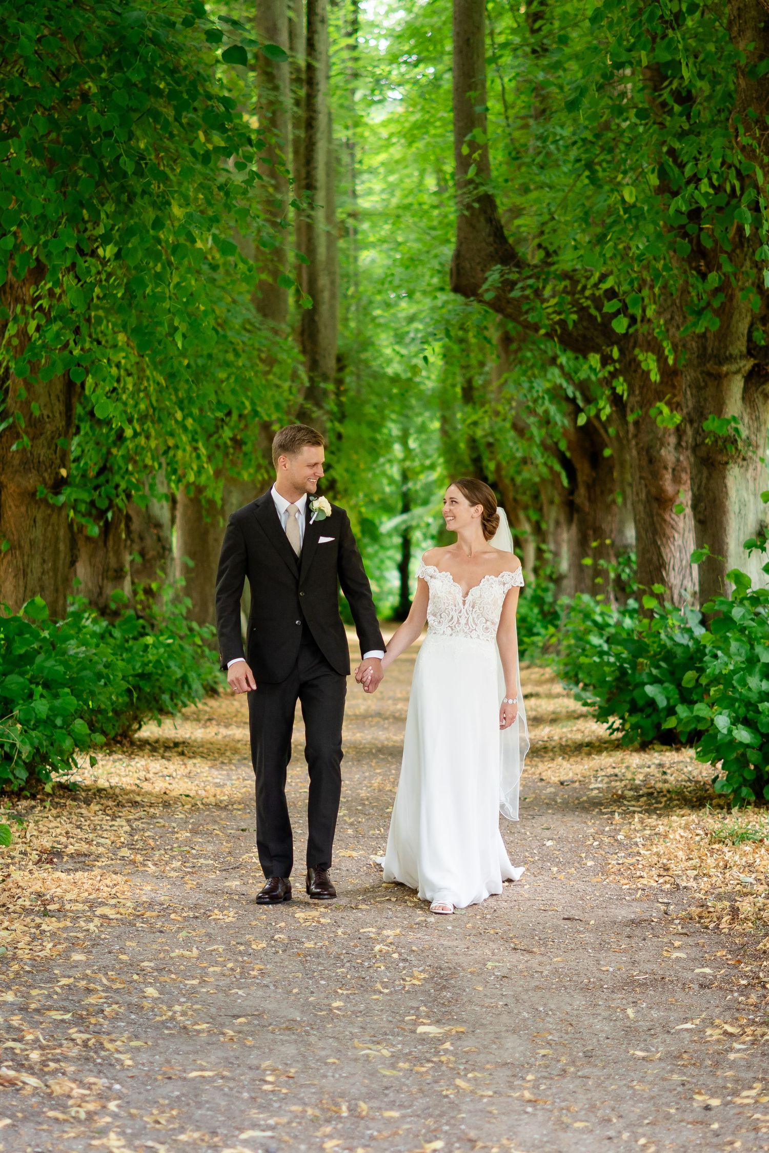 Bride and groom walk hand in hand down a tree-lined path in a lush green park on their wedding day.
