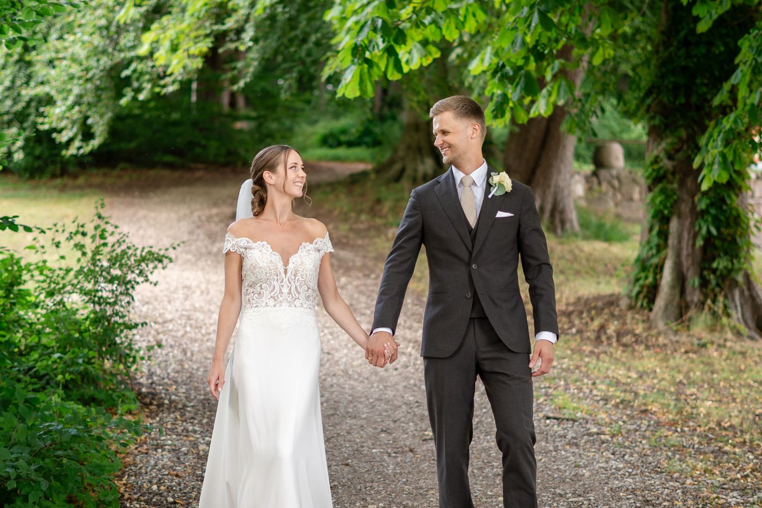 Bride in a white lace gown and groom in a dark suit hold hands while walking down a tree-lined gravel path.