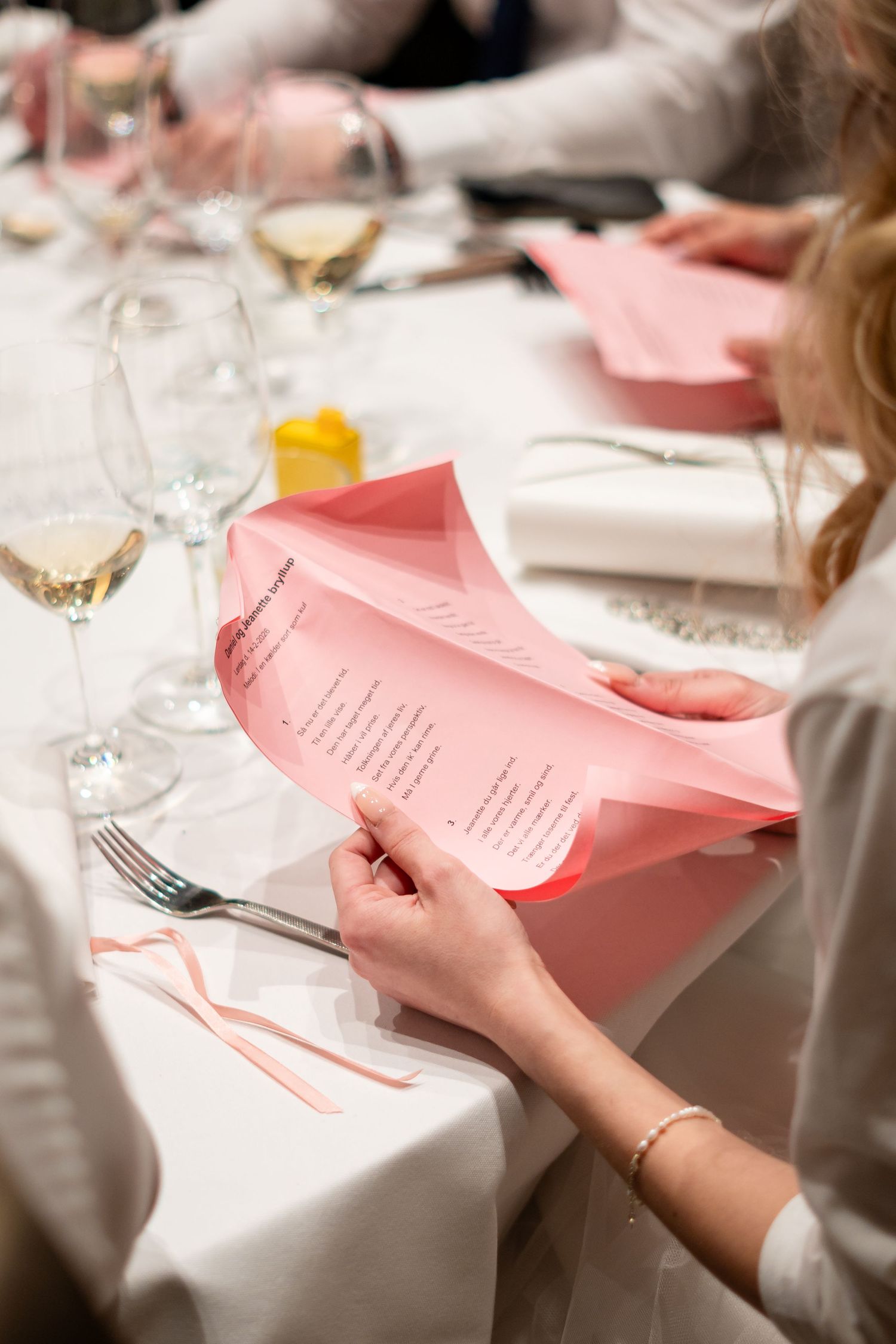 Guest reading pink menu card at formal table setting with wine glasses.