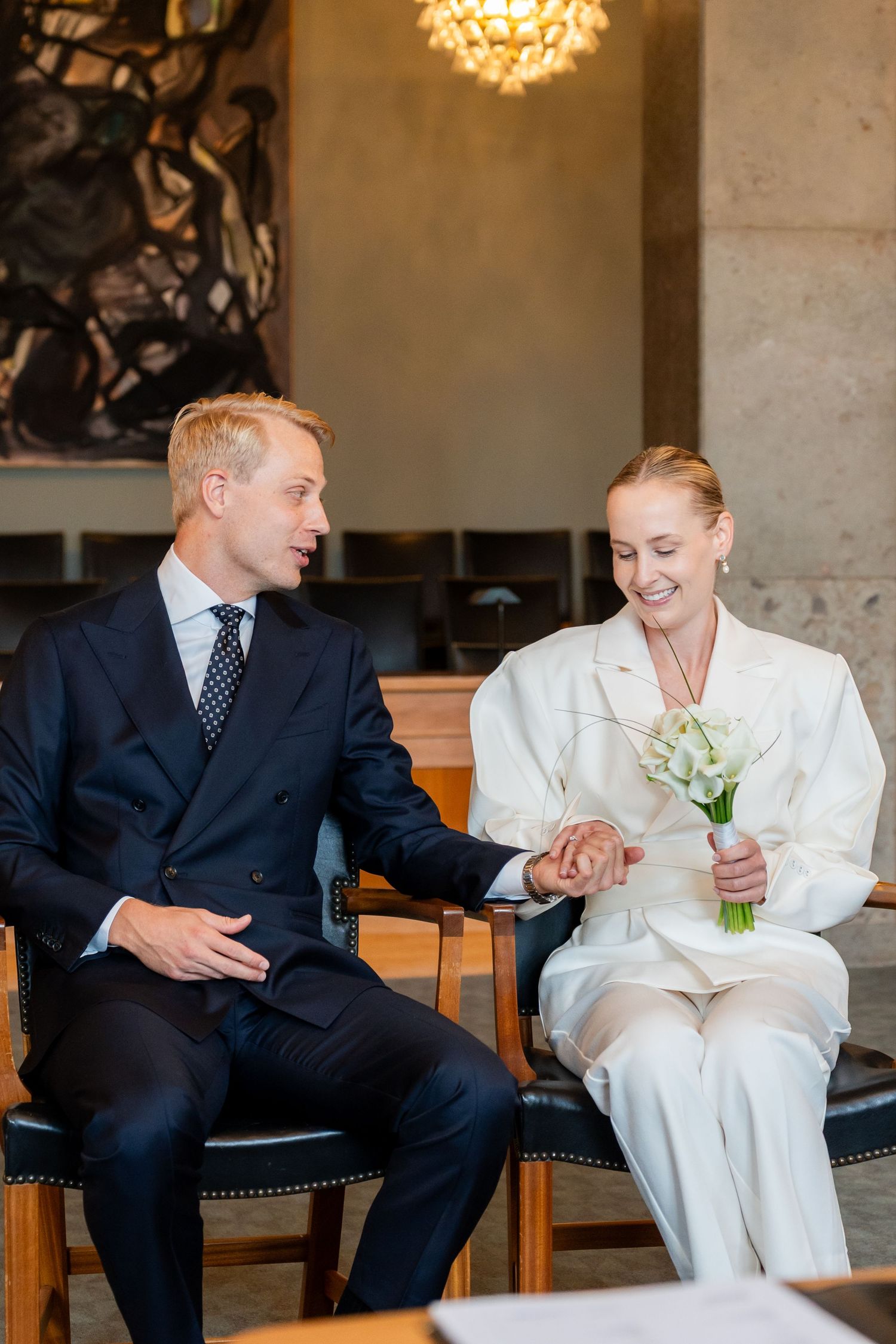 Bride in white suit holding calla lily bouquet sits with groom in navy suit, holding hands at elegant wedding ceremony.