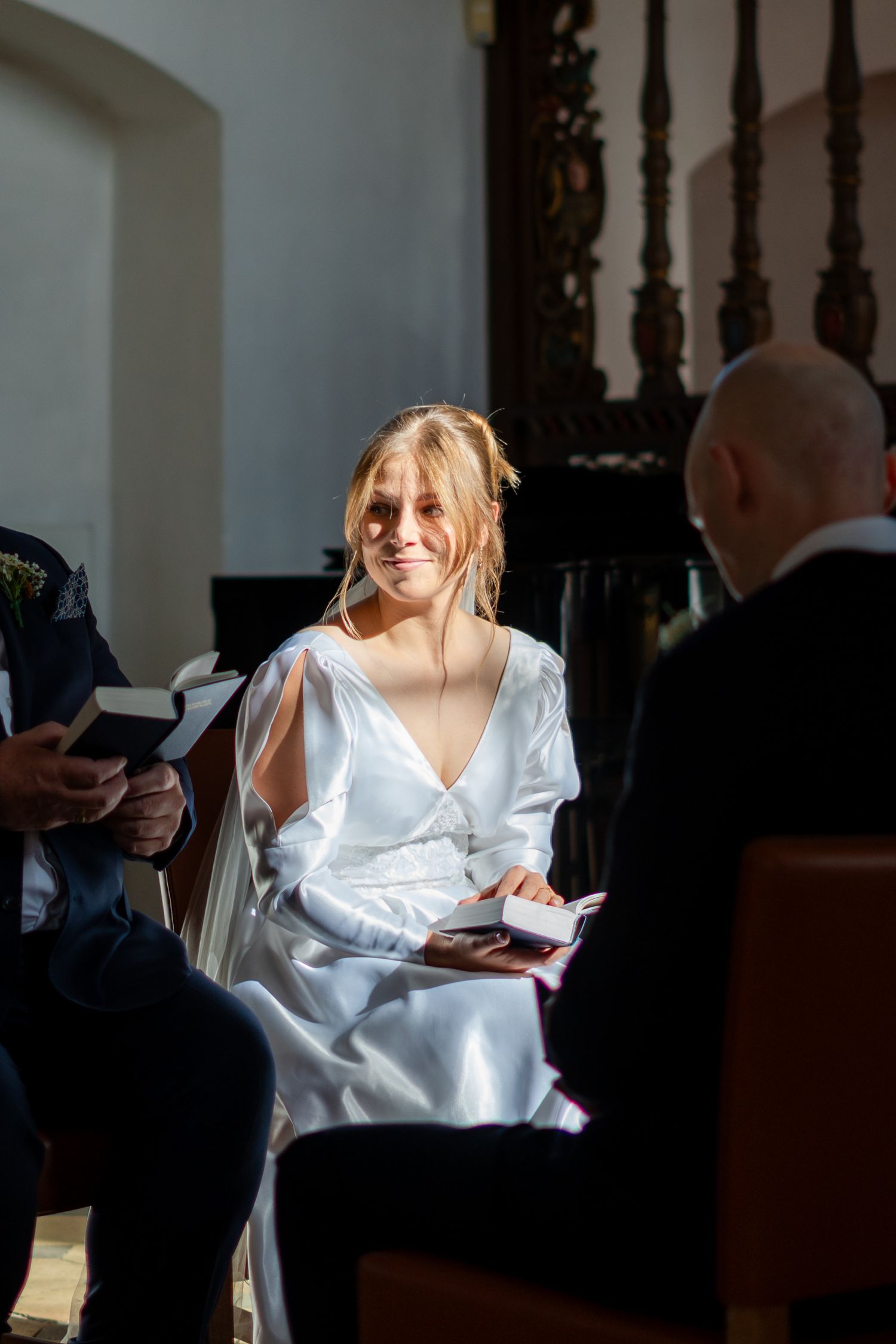 Bride in white satin dress smiling at groom during church wedding ceremony.