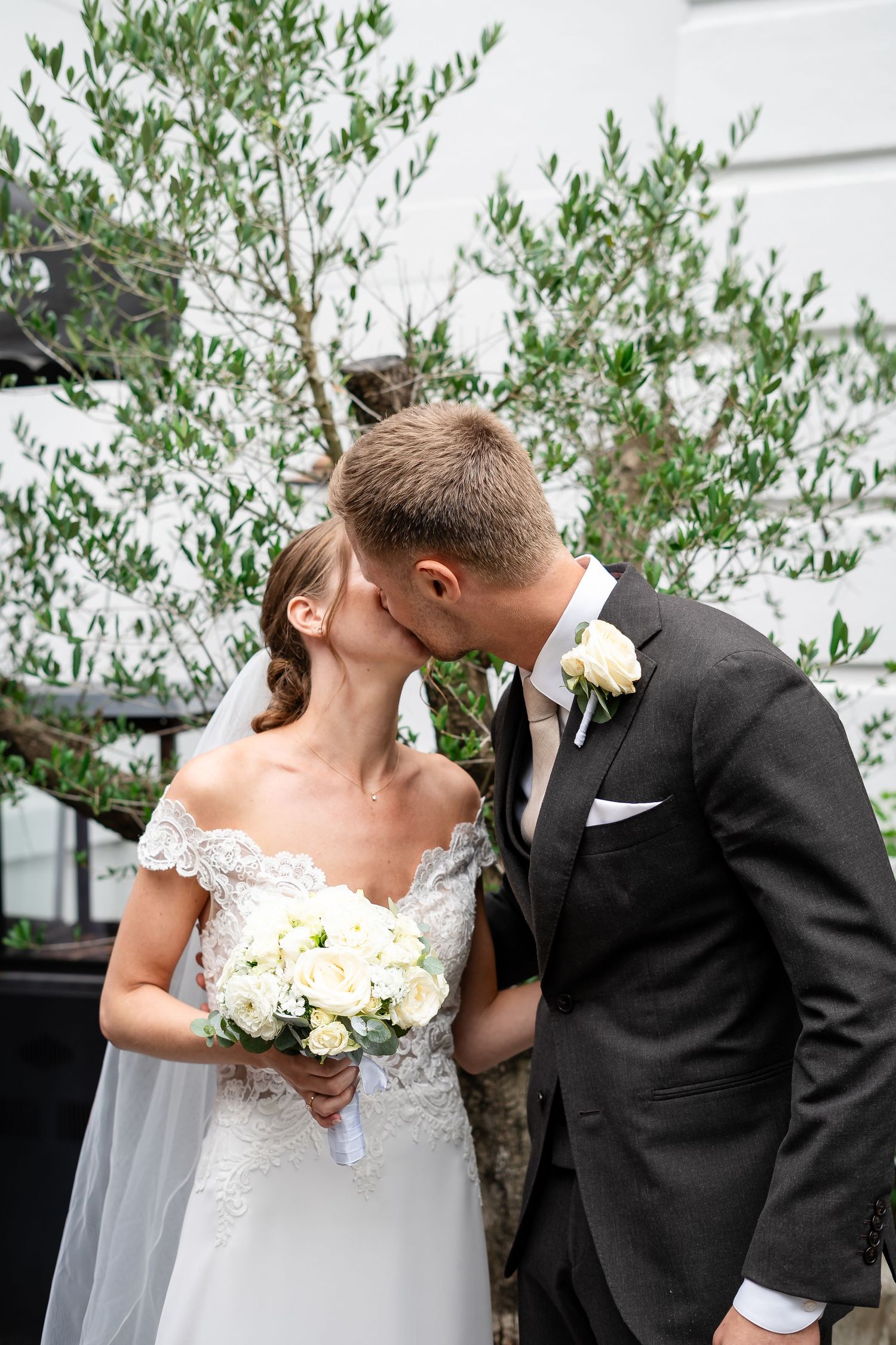 Bride and groom share a romantic kiss outdoors, bride holding white rose bouquet in lace off-shoulder gown with veil.