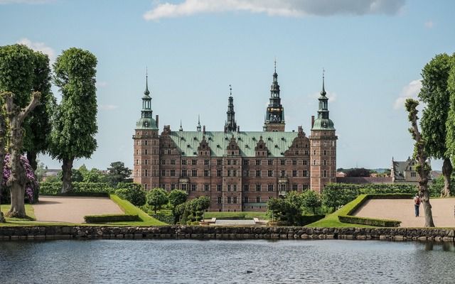 Panoramic view of Frederiksborg Castle with its spires and towers reflected in the lake on a sunny day.